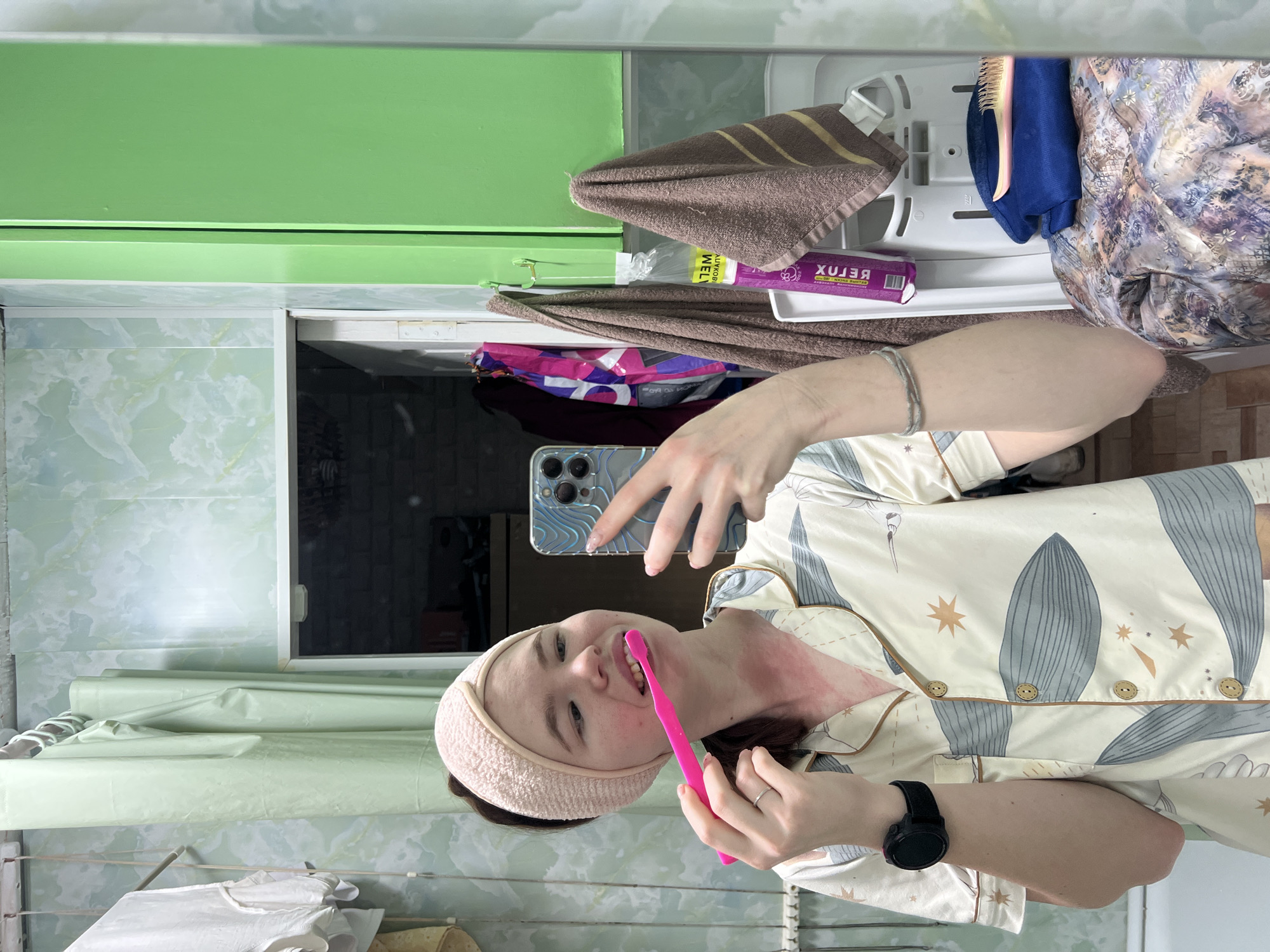A woman is in a bathroom, likely inside an apartment in Chaikovsky, Russia, taking a selfie while brushing her teeth with a pink toothbrush. She is wearing a pale peach headband and patterned pajamas. Her phone shows a reflection of the bathroom, including green cabinets and a mirror. The bathroom contains various items such as towels and personal care products on shelves. The walls have a light green marble-like pattern. A window is partially visible. The scene is domestically mundane.