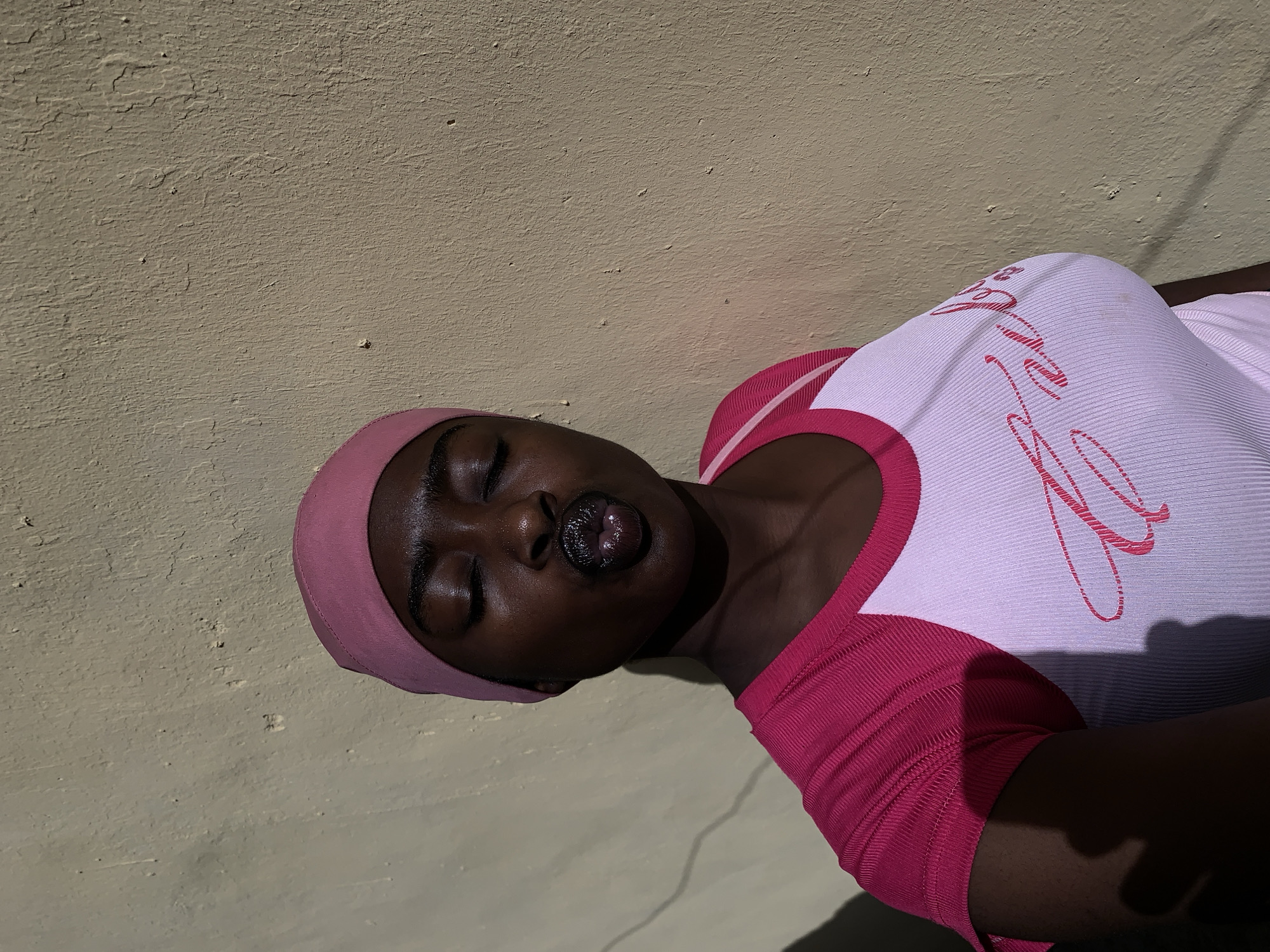 An image, originally rotated, features a dark-skinned person angled diagonally, with their head towards the upper left and body extending to the lower right. The person has their eyes closed and their glossy lips slightly parted, conveying a serene or relaxed expression. They wear a light pink head covering and a ribbed top. The top's main body is white with pink script text, which is partially obscured and illegible, while the collar and short sleeves are solid pink.
The background consists of a textured, light beige wall that shows signs of wear or peeling paint. Strong, harsh shadows fall across the person and the wall, indicating bright, direct sunlight, likely during midday or early afternoon, suggesting clear weather. These shadows create a dramatic contrast, emphasizing the textures of the wall and the ribbed fabric of the top. The overall mood is calm and tranquil, with the person appearing to be at rest or in a contemplative state. The scene suggests an outdoor setting, consistent with Rafin Par, Nigeria.