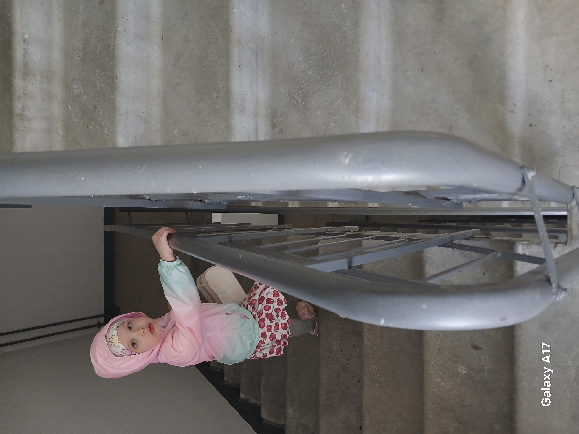 A photograph shows a small child clinging to a railing on a stairwell. The child, wearing a pink hooded jacket with light blue sleeves and a cherry-patterned skirt, hangs by one hand from the railing. The background consists of concrete steps and a painted wall. The location is not specified in the image and not definitively discernible from the provided visual information. The camera used to take the photo is a Galaxy A17, as indicated in the image.