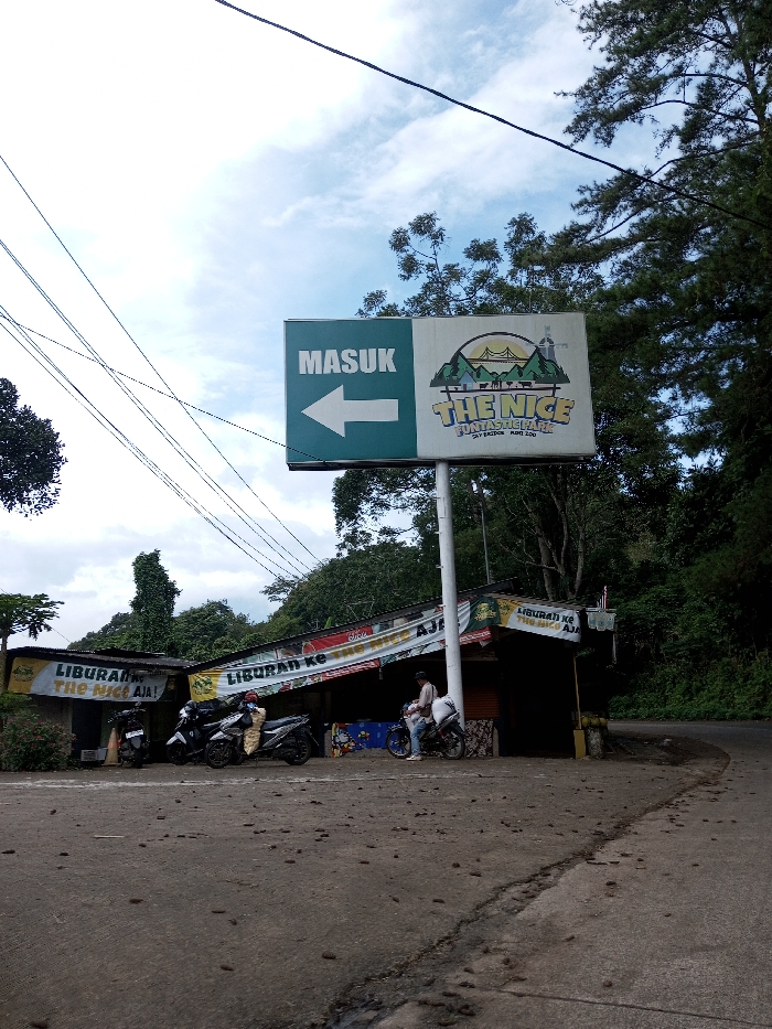 The image depicts an outdoor scene at what appears to be an entrance or roadside stop for an attraction. A prominent, large rectangular signboard, mounted on a pole, dominates the center. The left half of the sign is green with the white Indonesian text "MASUK" (Enter) and a large white left-pointing arrow. The right half is white, featuring a graphic logo of mountains, a bridge, and trees, with the text "THE NICE FUNTASTICK PARK" and "SKY BRIDGE MAY 200" below it. This signage strongly indicates an Indonesian location.
Below and to the left of the main sign, two contiguous, single-story structures with corrugated metal roofs are visible. These structures appear to be small shops or stalls, adorned with banners displaying the Indonesian text "LIBURAN ke The Nice Aja!" (Just holiday to The Nice!). Three motorcycles are parked in front of these structures on a paved surface. On the right side, an individual is seated on one of the motorcycles, facing away from the camera, wearing a light-colored top and pants. A second individual stands immediately adjacent to the motorcycle's right side, also facing away, dressed in a light top and dark pants. A bicycle leans against the rightmost structure.
The foreground consists of a dark, uneven asphalt or concrete surface with scattered debris and a noticeable crack running across it. The background and right side of the image are characterized by dense, lush green foliage and numerous tall trees, suggesting a hilly or mountainous environment. Utility lines crisscross the upper left portion of the frame against a partly cloudy sky.