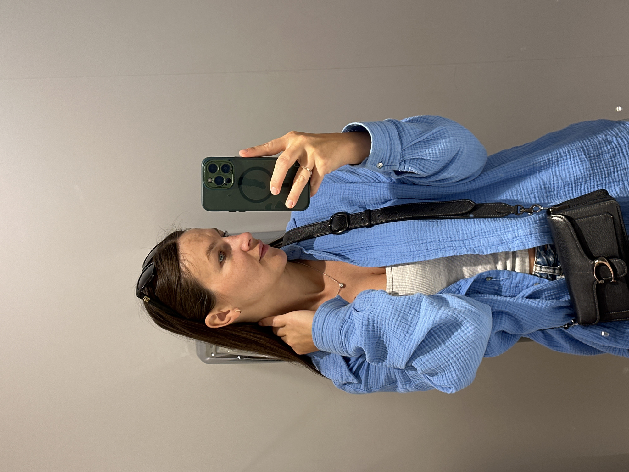 A woman takes a selfie in an indoor location in the city of Unknown, Unknown. She holds a green smartphone in her right hand, showcasing a wedding ring on her finger. She has brown hair and is wearing a blue crinkle shirt. The shirt is accessorized with a black crossbody bag. Sunglasses are perched on top of her head. The backdrop appears to be a plain, light-colored wall.