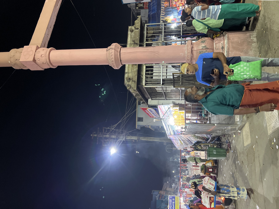 The image captures a vibrant nighttime street scene in Palani, India, likely near a temple or market area. A large, light pink/beige architectural structure, possibly a carved pillar or part of a gateway, stands prominently on the left, adorned with decorative elements.
In the foreground, two men are engaged in conversation. One man, on the right, wears a dark blue and light blue collared shirt, light-colored trousers, and a yellow skullcap. He holds a smartphone in one hand and a bright green carry bag in the other. Facing him, on the left, is an older man with a beard and a tilak on his forehead, dressed in a dark green shirt and an orange/red traditional dhoti or lungi. He also wears bracelets on his wrist.
The background reveals a bustling environment with numerous people, many dressed in traditional Indian attire. Several vendor stalls are visible, brightly lit and displaying various goods, creating a lively atmosphere. A bright street light illuminates the right side of the image, casting a strong flare. Overhead power lines crisscross the dark night sky.
Visible text includes "GATE-3" on a yellow sign near the bottom right, and a smaller sign bearing the number "26263." The overall scene depicts a typical busy Indian street at night, filled with activity and commerce.