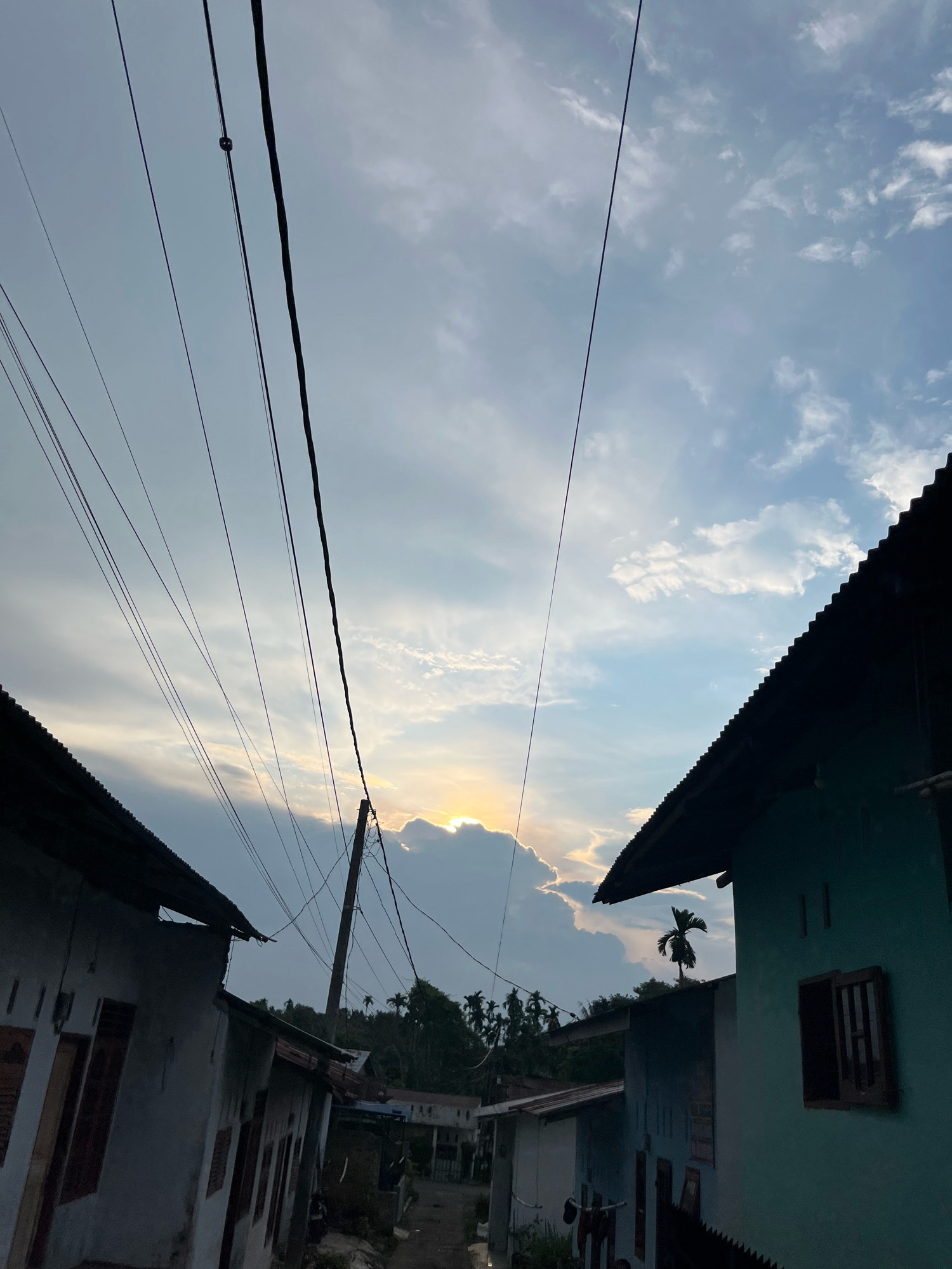 This is a vertically oriented, low-angle shot capturing a street view in Patumbak Kampung, Indonesia, during what appears to be sunset. The foreground is dominated by the dark silhouettes of the corrugated metal roofs of two houses, one on the left with a white facade and wooden window frames, and another on the right painted a muted teal with a visible wooden-framed window. A series of dark electrical wires crisscross the sky, originating from a wooden utility pole visible between the two houses and stretching towards the upper left of the frame. The sky above is a mix of blue and grey clouds, with the sun peeking through a large, bright cloud formation towards the center-right, casting a warm glow. In the background, a line of palm trees and other tropical foliage suggests a rural or semi-rural setting. The street between the houses is narrow and unpaved, with some indistinct structures and vegetation visible further down. No people are present in the frame, and the scene is static, focusing on the architectural elements and the dramatic sky.