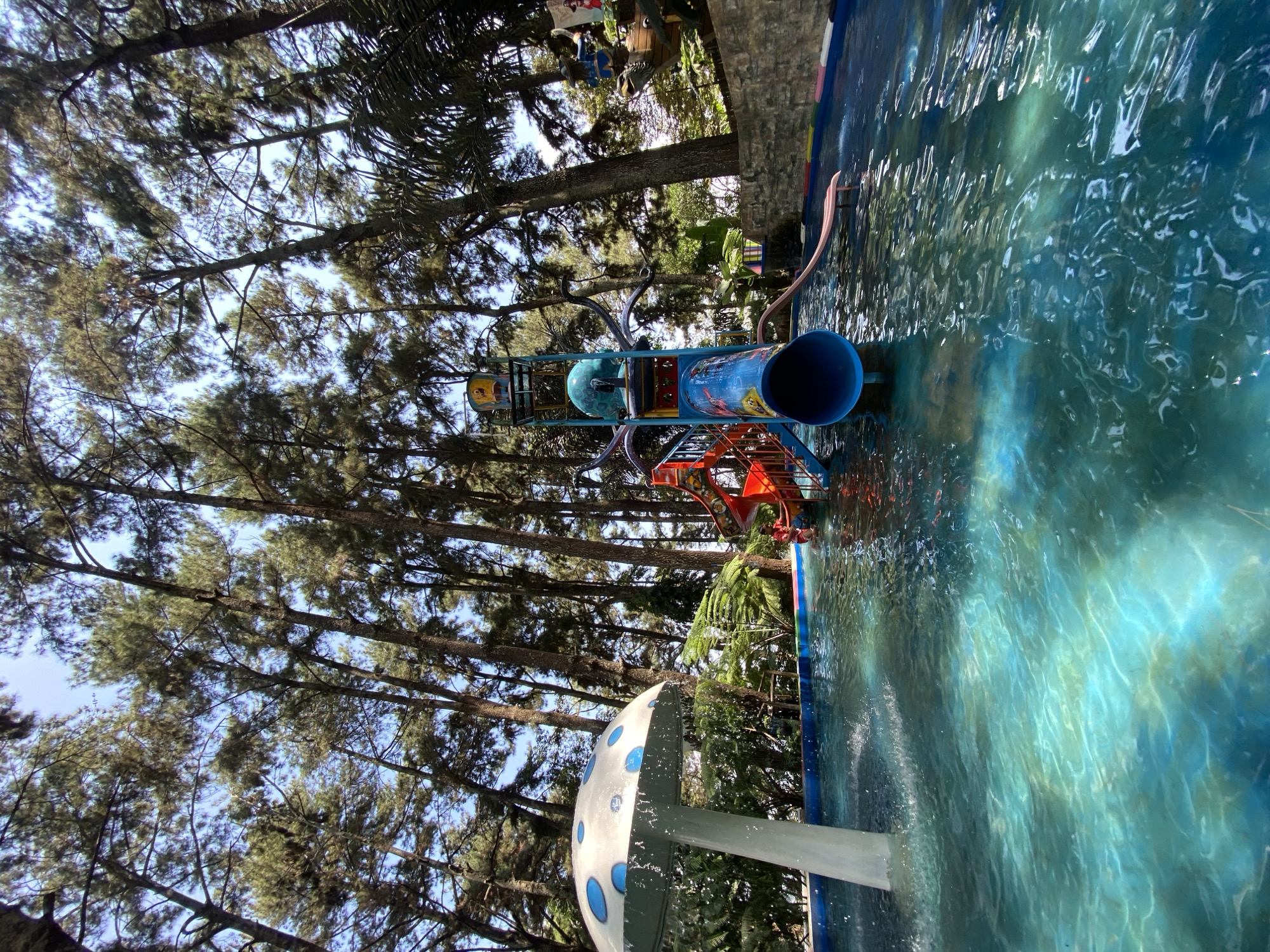 In Kuwukan, Indonesia, a water park features a wading pool surrounded by tall trees. A mushroom-shaped water feature with blue polka dots stands on the left side of the pool, with water spraying from it. In the center, a multi-colored water slide structure, primarily blue, red, and yellow, is positioned. A person in red swimwear is visible near the bottom of the slide, appearing to be in the water. The pool's water is turquoise, with sunlight creating ripples and reflections on its surface. A stone wall borders the background of the scene, with lush greenery and palm trees visible behind it. The sky is clear and blue, with the sun illuminating the area.