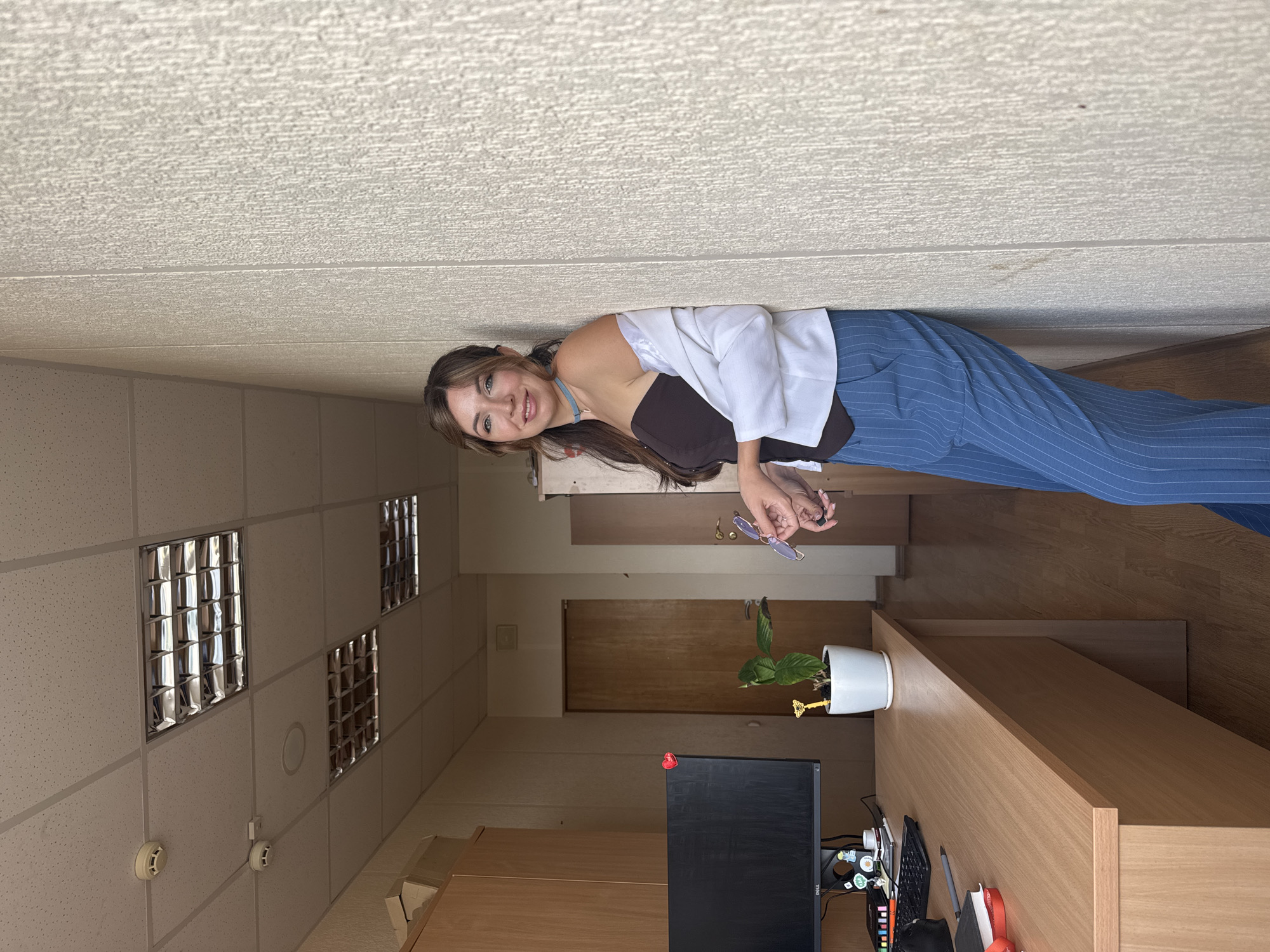 A young woman with long brown hair, wearing a brown strapless top, a white open blazer, and blue pinstripe pants, is leaning against a textured wall in an office. She is holding a pair of glasses in her right hand. The office features a wooden desk with a computer monitor, keyboard, a potted plant, and various office supplies. The ceiling has fluorescent lighting panels and smoke detectors. The floor is made of wood. In the background, there are wooden doors and cabinets. The image appears to be taken in Kryvyi Rih, Ukraine.