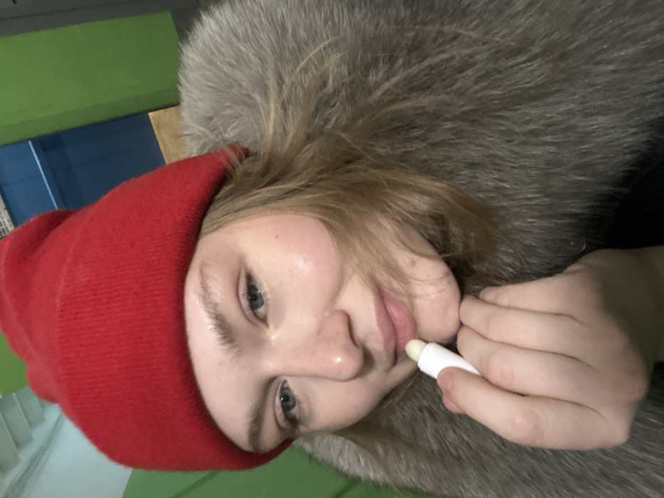 A fair-skinned woman wearing a red beanie and a fur-trimmed hood applies lip balm. Her blue eyes look toward the camera, and her lips are slightly parted. The background includes a green surface and a glimpse of blue, possibly a wall. The image is taken at an angle, with the woman's face rotated toward the viewer. Her hand holds a white tube of lip balm, which she applies to her lips.