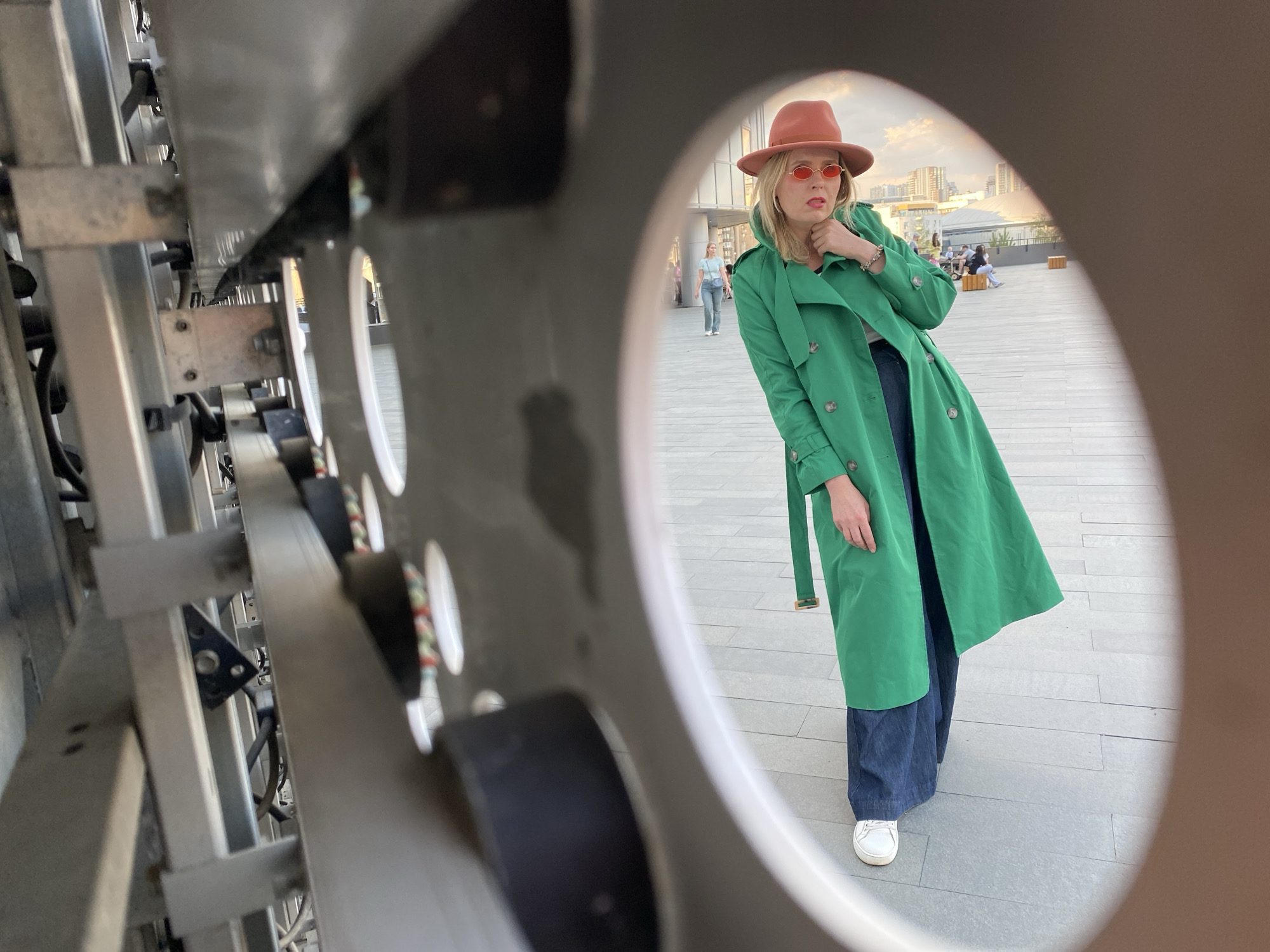 A woman with blonde hair, wearing a brown hat, red oval sunglasses, a green trench coat, dark blue pants, and white sneakers, is viewed through a circular opening of what appears to be a metal sculpture or architectural feature. The woman is looking off to the left. The background includes a paved area, other people, and buildings. The location is Yekaterinburg, Russia.