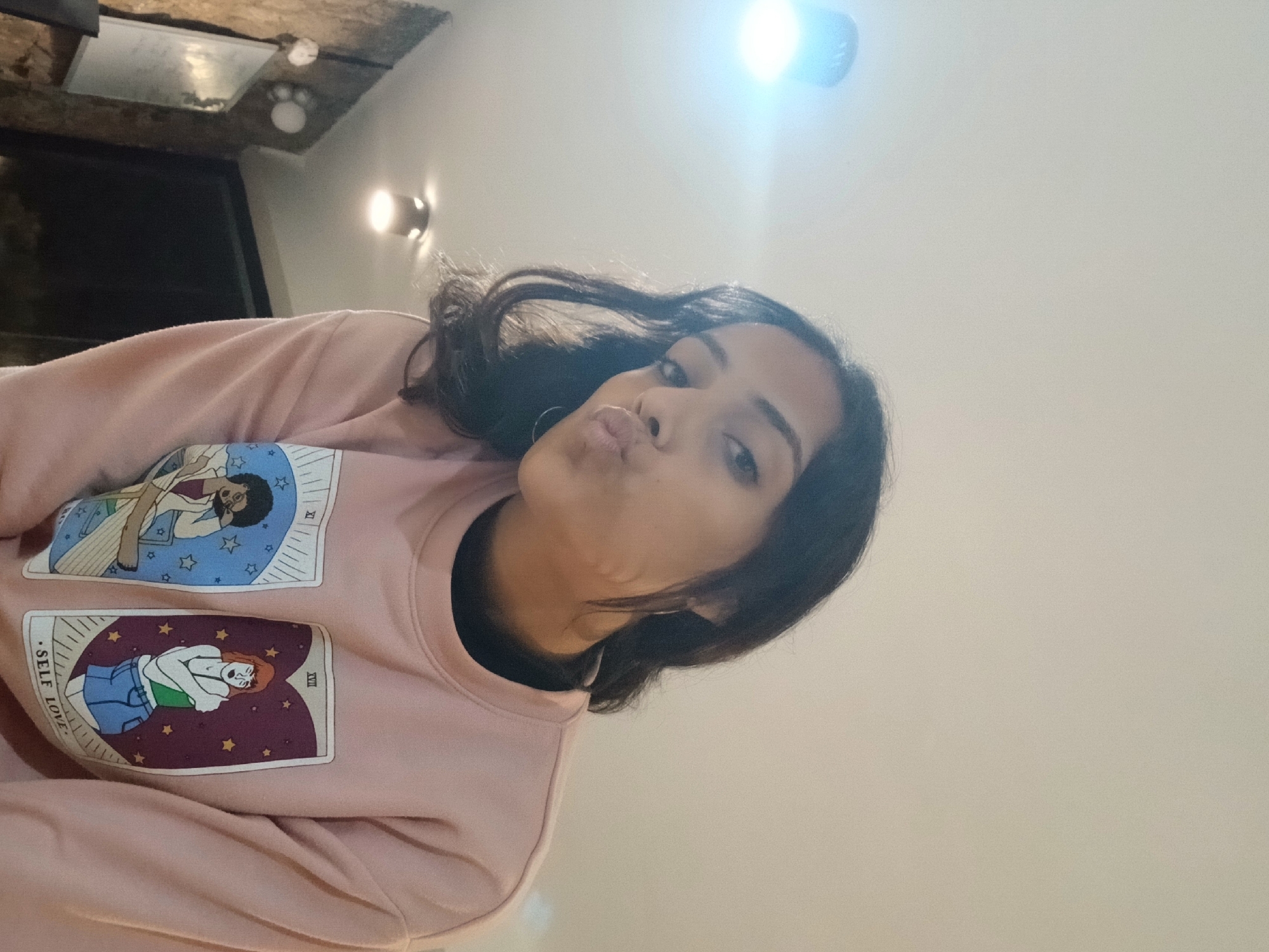 A young woman with dark hair and hoop earrings is taking a selfie. She is wearing a pink sweatshirt with two tarot card designs on the front, one labeled "Self Love." The background appears to be an indoor setting with a light fixture on the ceiling and a wooden-paneled wall. There is no indication of the location being Playa El Agua, Venezuela.