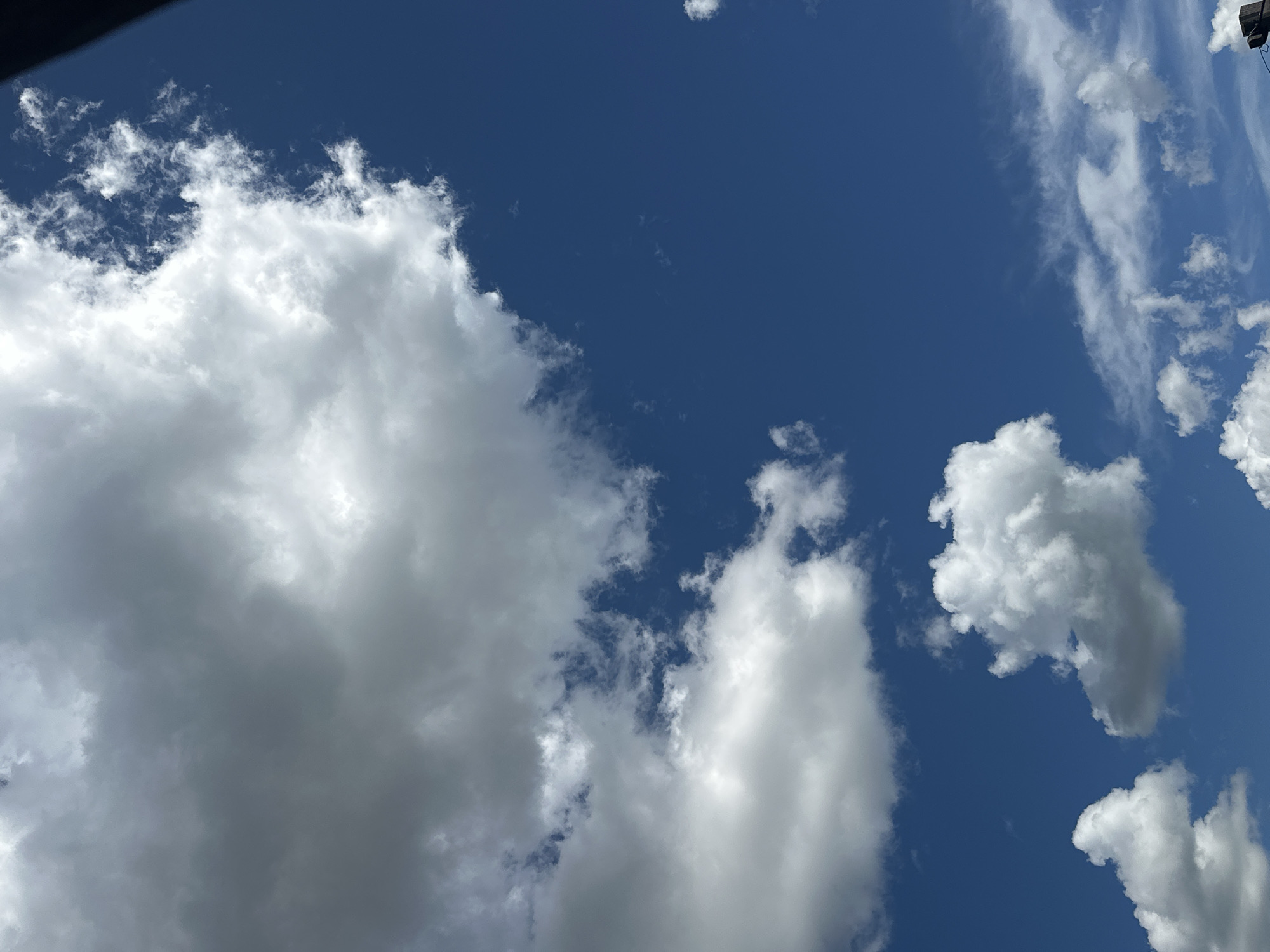 The image shows a bright blue sky with a variety of clouds. In the foreground, there are several large, fluffy cumulus clouds, some of which are lit brightly by the sun. In the background, there are thinner, wispy cirrus clouds. A dark, vertical object, possibly a power pole or antenna, is visible in the lower right corner of the image. The scene is likely outdoors, under an open sky. This image does not provide any specific visual information to determine its location in the city of Tobyl, Kazakhstan.