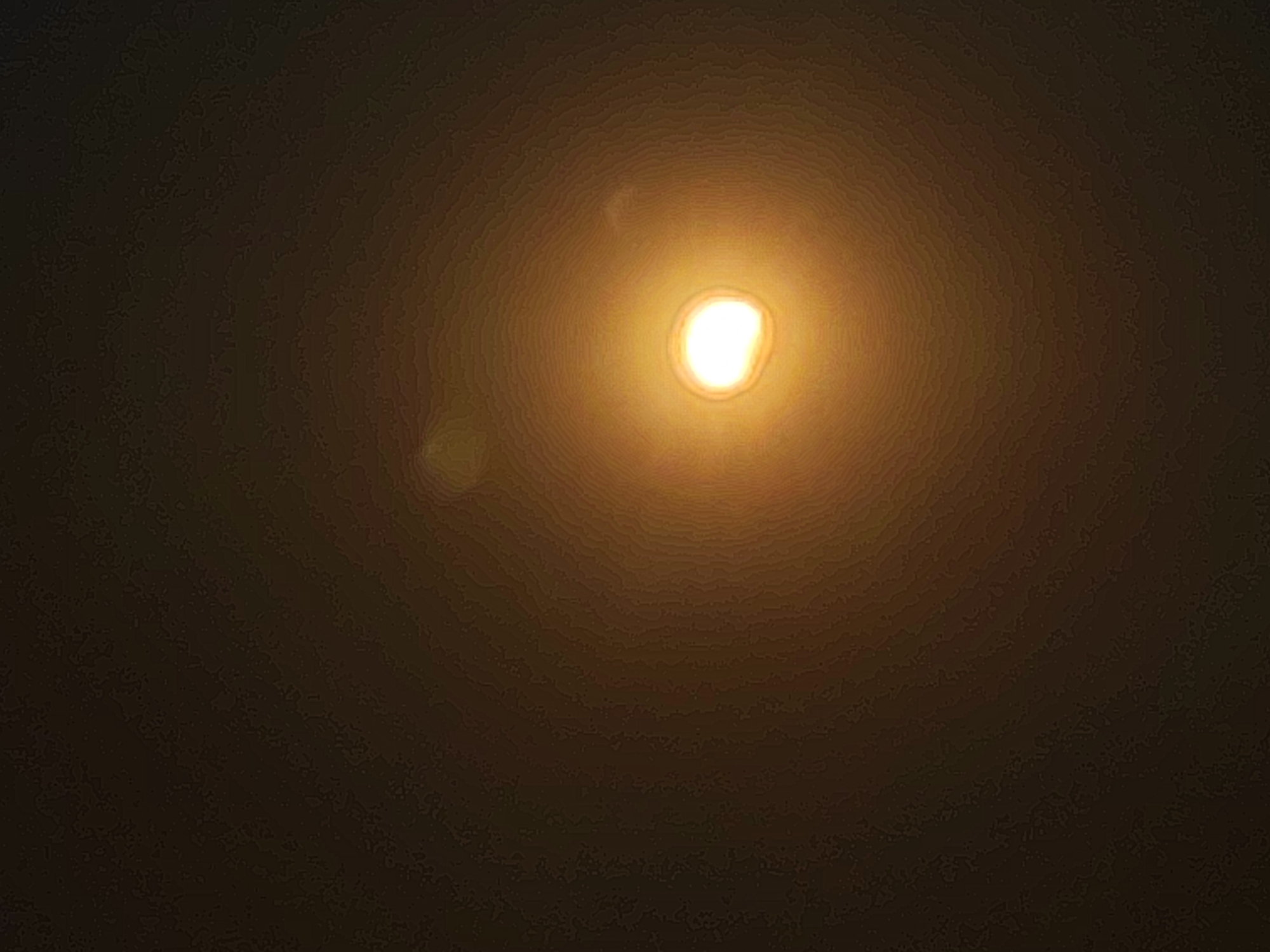 The image displays a bright, circular light source, likely the sun, positioned centrally within a dark, textured background. Concentric rings of light emanate from the central source, creating a halo effect. There are faint lens flares visible as smaller, amorphous light shapes scattered around the primary light source. The overall impression is of looking directly at a strong light through a hazy or atmospheric medium. The image is devoid of any identifiable features of the city of Autillo de Campos, Spain, or any people or specific objects.