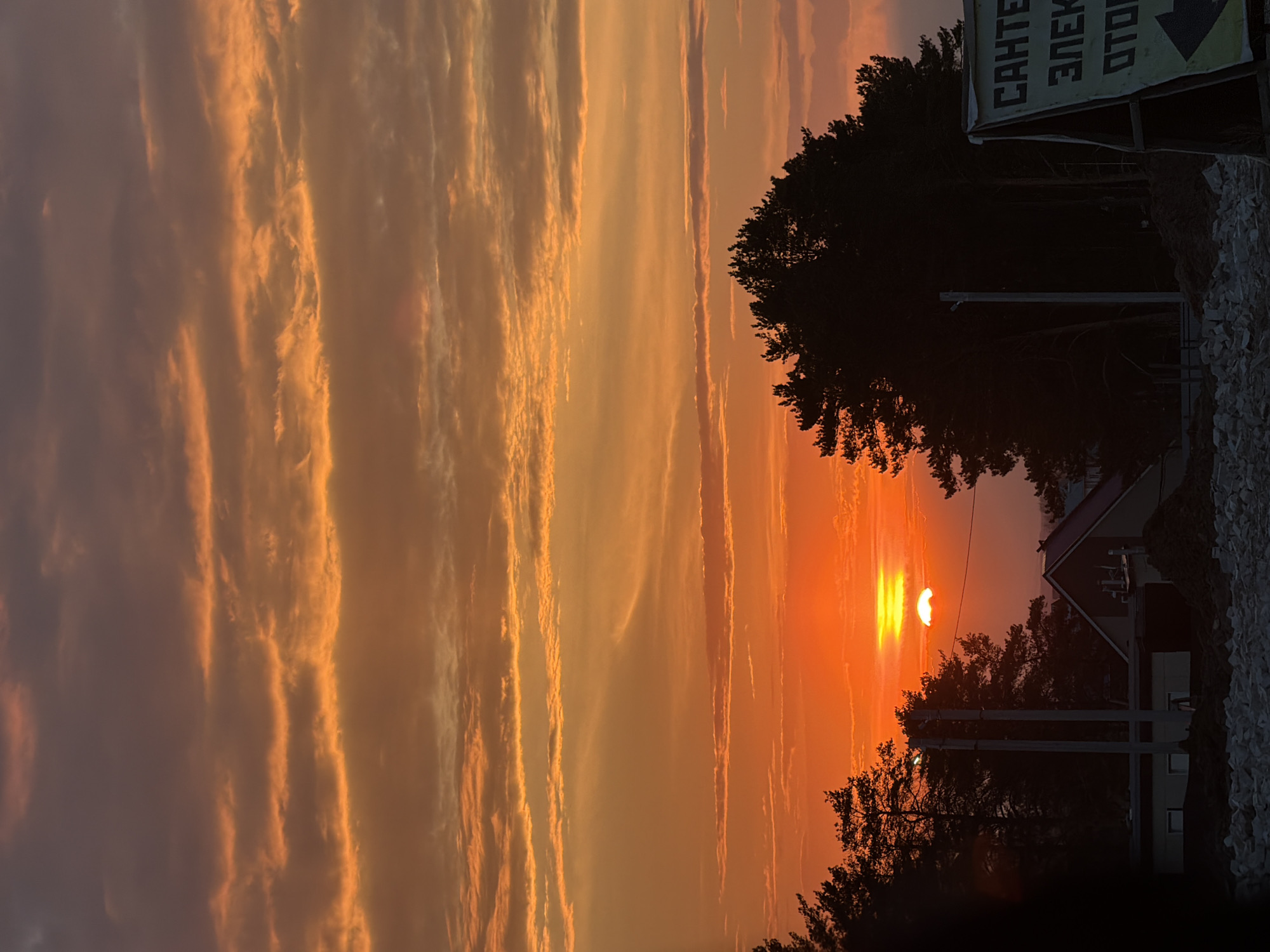 The image captures a sunset in Chaikovsky, Russia. The sky is filled with orange and yellow clouds, creating a vibrant backdrop. The sun is partially visible, appearing as a bright orange disc, partially obscured by an unknown object, possibly the moon. Silhouetted trees are visible in the foreground, along with a building with a pointed roof. A sign with Cyrillic text is present on the right side of the frame. The scene suggests a peaceful, twilight environment.