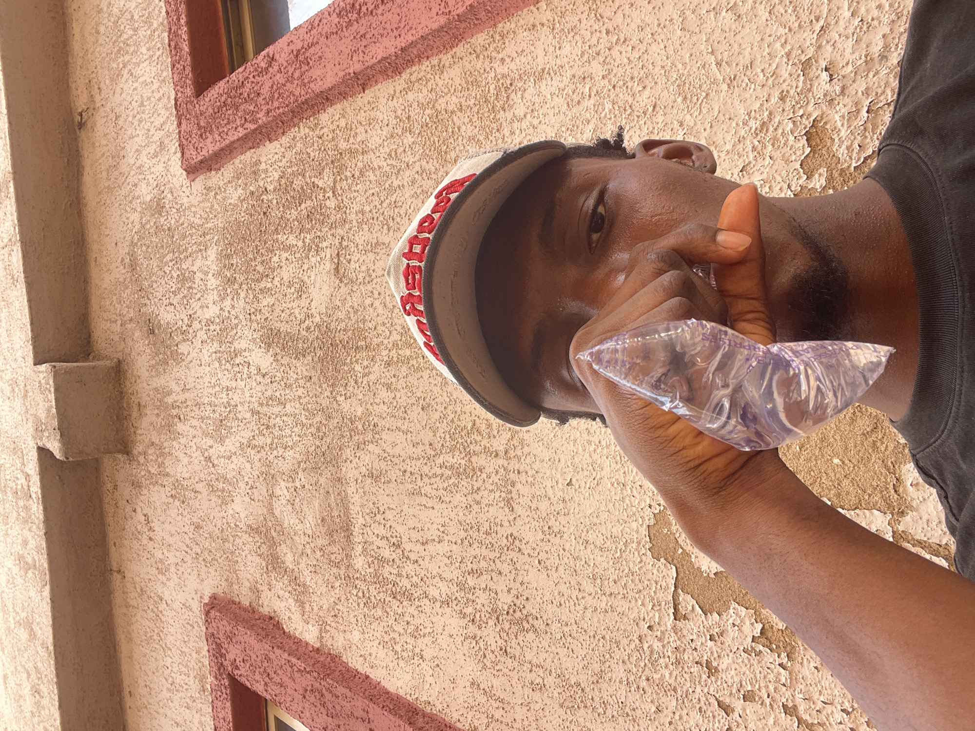 The image, rotated vertically, captures an upward-angled shot of a young Black male drinking from a clear plastic sachet. He is seen from the chest up, wearing a light-colored baseball cap with red embroidery and a dark t-shirt. His face and neck are visibly sweaty, indicating warm weather. His right hand holds the sachet to his mouth as he drinks.
The background is an exterior building wall, featuring a textured, stucco-like finish in a light beige or tan color. Patches of peeling paint are visible on the wall, suggesting age or environmental wear. A reddish-brown painted window frame is partially visible in the upper right, and a vertical architectural element, possibly a beam or corner, runs along the left side of the frame. The bright, direct lighting suggests it is daytime.
Visible text on the brim of the cap clearly spells "DRINK" in red lettering. On the plastic sachet, faint text includes parts that resemble "APPR" and "SAPE," potentially indicating "APPROVED" and "SAPELE WATER," a common type of packaged water in Nigeria, aligning with the given location context of Lokoja.