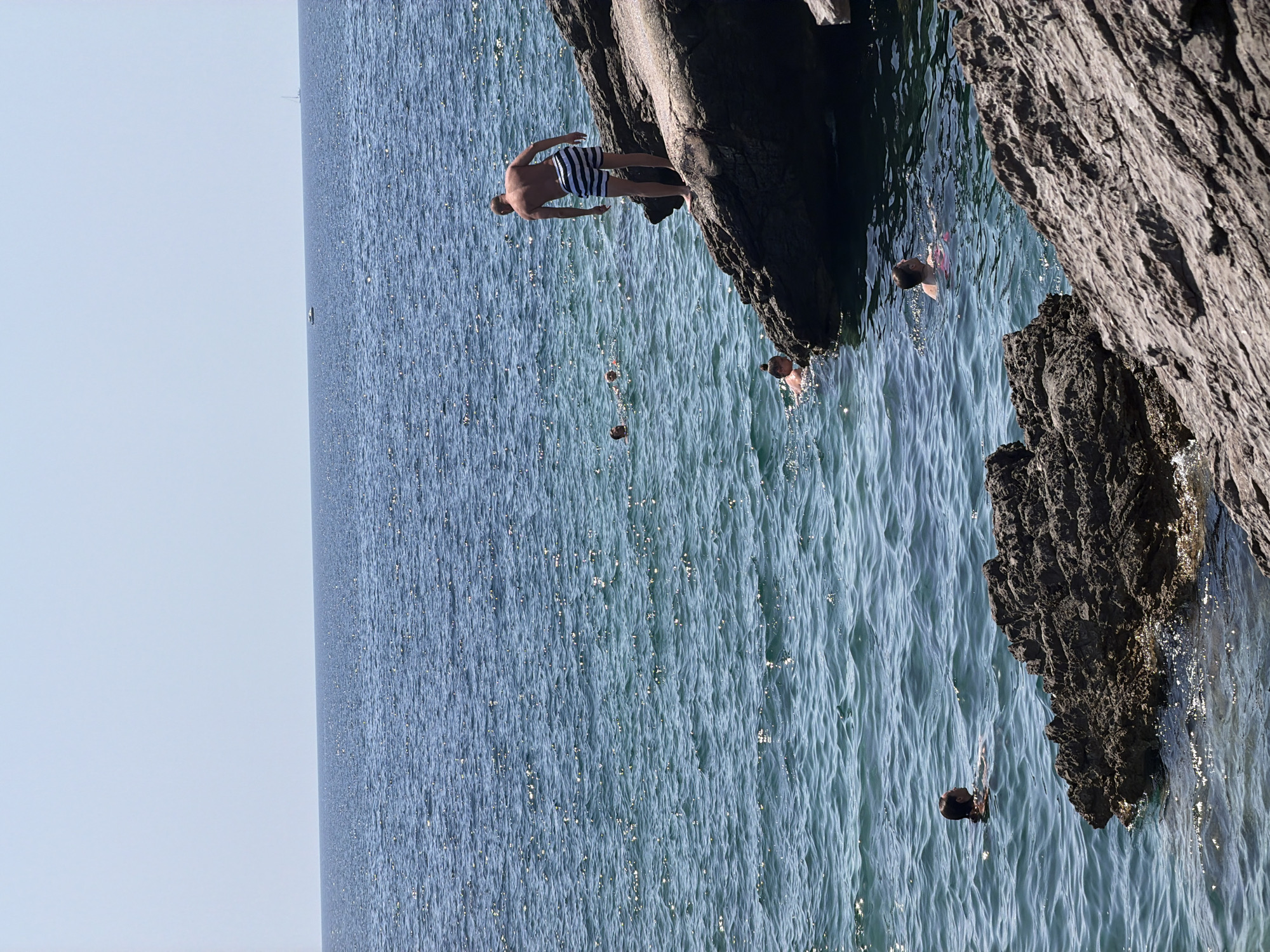 In Bigova, Montenegro, several people are swimming in the clear blue Adriatic Sea. A man in striped swim trunks stands on a large rock overlooking the water, appearing to be about to jump in. In the water, a woman with dark hair tied back swims towards the shore, while two other individuals are partially visible in the background. The sun glints off the rippling surface of the water, and rocky outcrops line the coastline. Two boats are visible in the distance on the horizon.