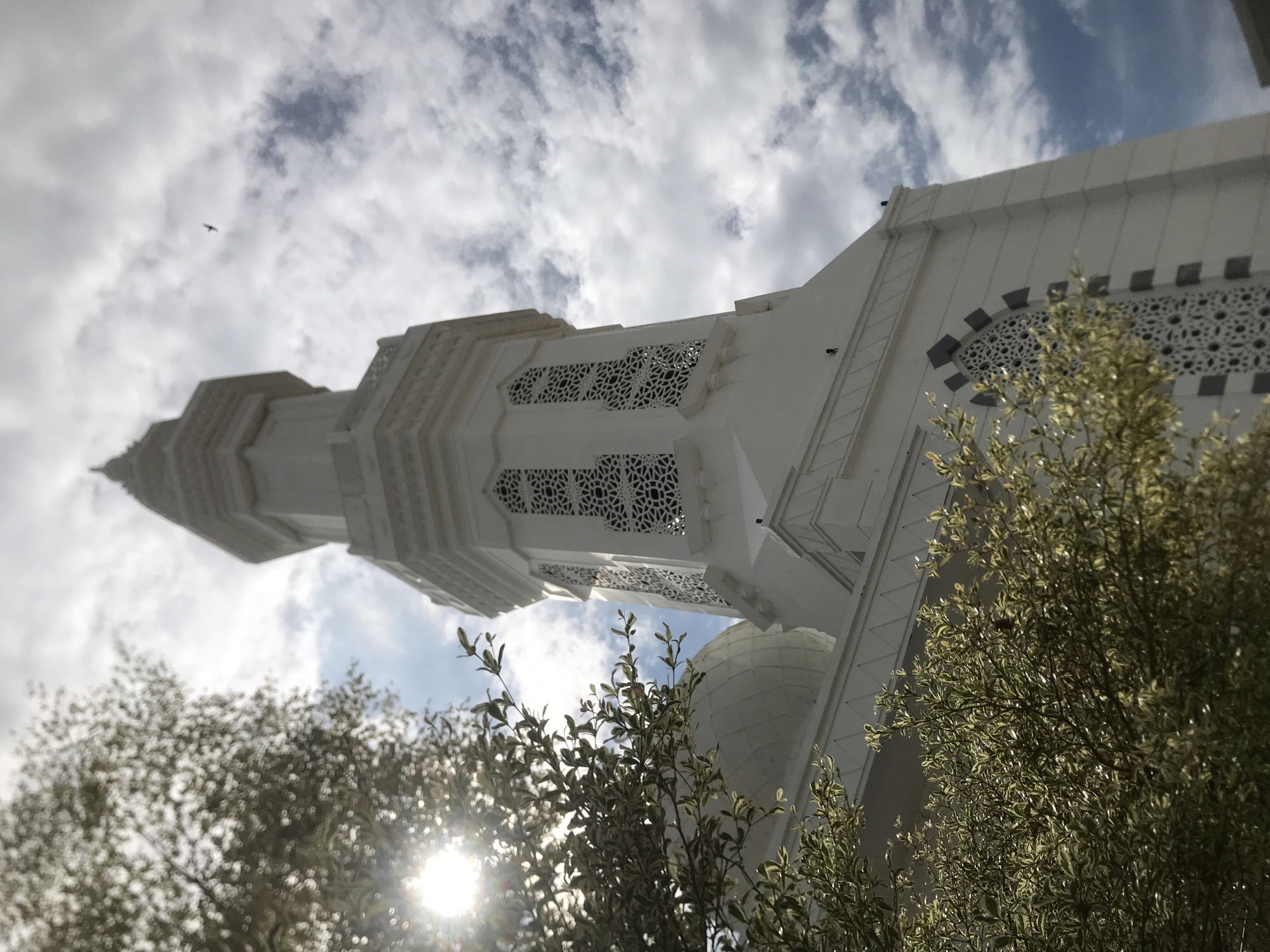 A low-angle shot shows a white building with geometric patterns under a cloudy sky, in Meunasahaluedrienlhoksukon Barat, Indonesia. In the lower portion of the frame, the sun shines through trees. A bird flies in the upper left portion of the frame.