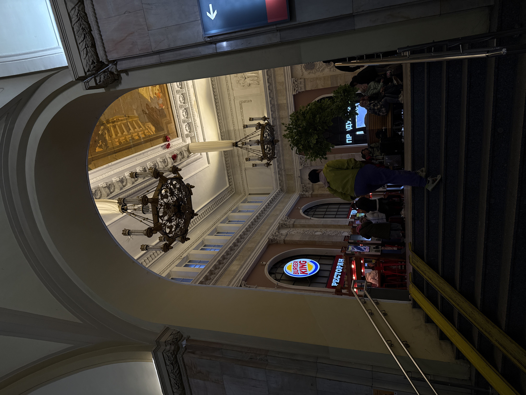 A low-angle shot looks through an arched doorway toward an ornate interior space. The foreground shows sections of arches and architectural detailing. In the mid-ground, the view opens to reveal a high-ceilinged area with classical architectural elements, including murals, columns, and large chandeliers. Visible beyond the architectural details are windows and a Burger King restaurant sign in Russian. People are situated on stairs to the right. An illuminated directional sign with a downward-pointing arrow is positioned above the staircase. The image suggests a location within the city of Saint Petersburg, Russia.