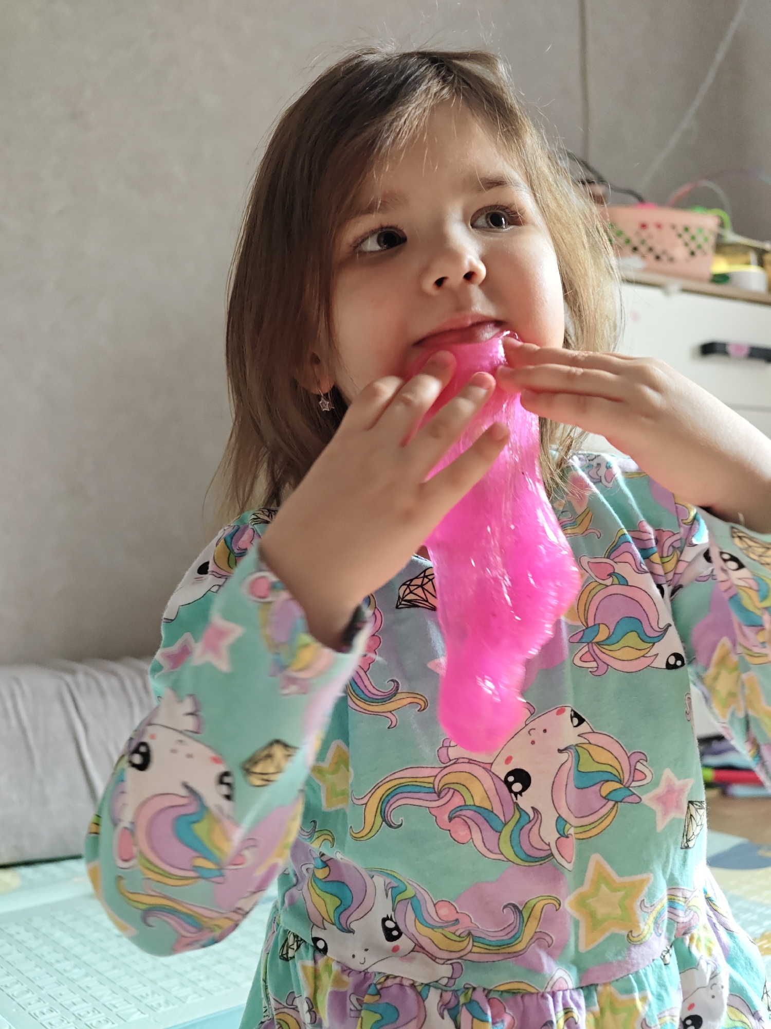 An indoor photograph, likely taken in a residential setting within the city of Chaikovsky, Russia. The central subject is a young Caucasian girl, appearing to be a toddler or preschooler, with shoulder-length light brown hair. She is wearing a long-sleeved light teal-colored top or dress patterned with white unicorns, rainbow manes/tails, colorful stars, and black outline diamonds. Small, gold-colored star-shaped hoop earrings are visible in her ears.
The girl's hands are actively engaged with a substantial quantity of bright pink, translucent, glossy slime, which she is stretching downwards from her lower lip and chin area. Her mouth is slightly open where the slime originates, and her gaze is directed upwards and slightly to the right of the frame.
The background on the left side features a light-colored, textured wall above a section of a white or light-grey upholstered furniture piece with horizontal ridges. On the right, partially visible elements include a pink plastic basket with a grid pattern and a segment of white furniture with a dark handle. The lower foreground shows a colorful play mat with geometric patterns, including a light blue-green section.