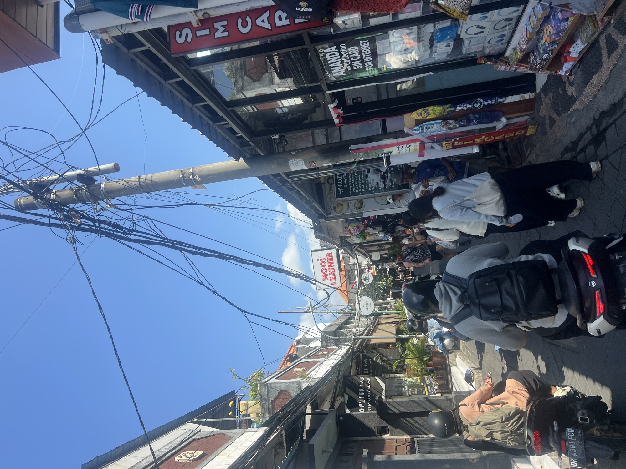 In Kuta, Indonesia, a narrow street is depicted with a bright blue sky above and a tangled mess of overhead electrical wires. The street is lined with shops on both sides, displaying various goods and signage. On the left, a shop labeled "OPTIMES TWO" is visible, alongside a brick building with decorative elements. The right side features shops with large windows, including one with a prominent red sign that reads "SIMCARD" and another with signage for "AMANDA SIM CARD FOR INTERNET". Various merchandise is displayed, including what appears to be headphones and other electronic items.
Several people are present, with a strong focus on individuals riding motorbikes. Two motorbikes are prominently in the foreground, with riders wearing helmets. The rider on the left wears a peach-colored top and a backpack. The rider on the right wears a grey hoodie and a black backpack. Other people are seen walking in the background, some appearing to be tourists or shoppers browsing the shops. The street is paved with stone or brick. The overall impression is of a busy commercial street in a tropical urban environment.