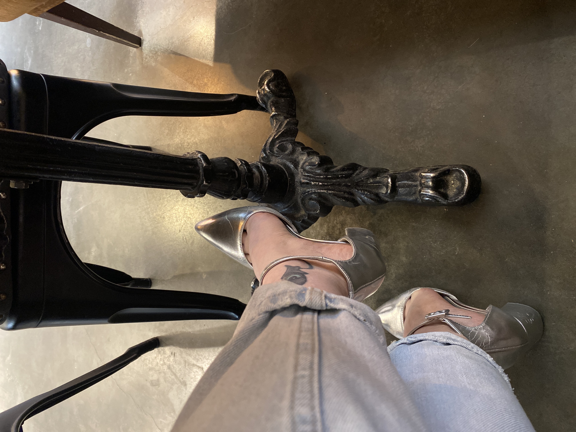 The image shows a top-down view of a person sitting at a table, likely in a restaurant or cafe. The person is wearing light-wash jeans and silver, pointed-toe heeled shoes with ankle straps. The right ankle has a small tattoo. The table has a black, decorative cast-iron base. The floor appears to be a neutral-colored, possibly concrete or tile surface. There is no context to determine location in Yekaterinburg.