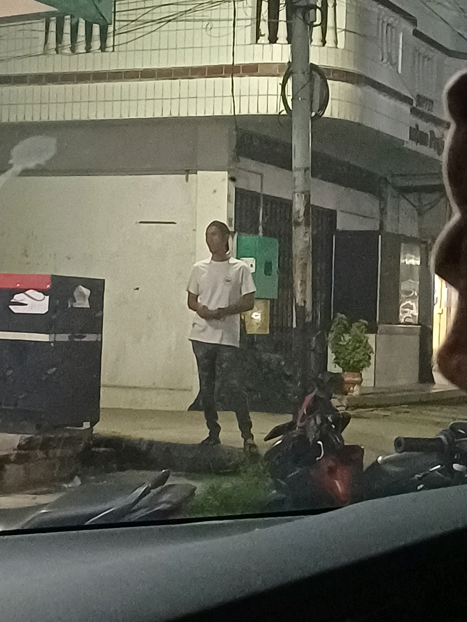In Dumai City, Indonesia, a person stands in front of a building, partially obscured by a dark vehicle in the foreground. The individual, dressed in a white t-shirt and dark pants, is positioned to the right of a dark trash receptacle with red and white markings. The building behind features a tiled facade with horizontal brown bands. A utility pole with wires stands near the person. A potted plant is visible to the right of the building entrance. The scene appears to be an outdoor urban environment, possibly at night or in low light conditions.