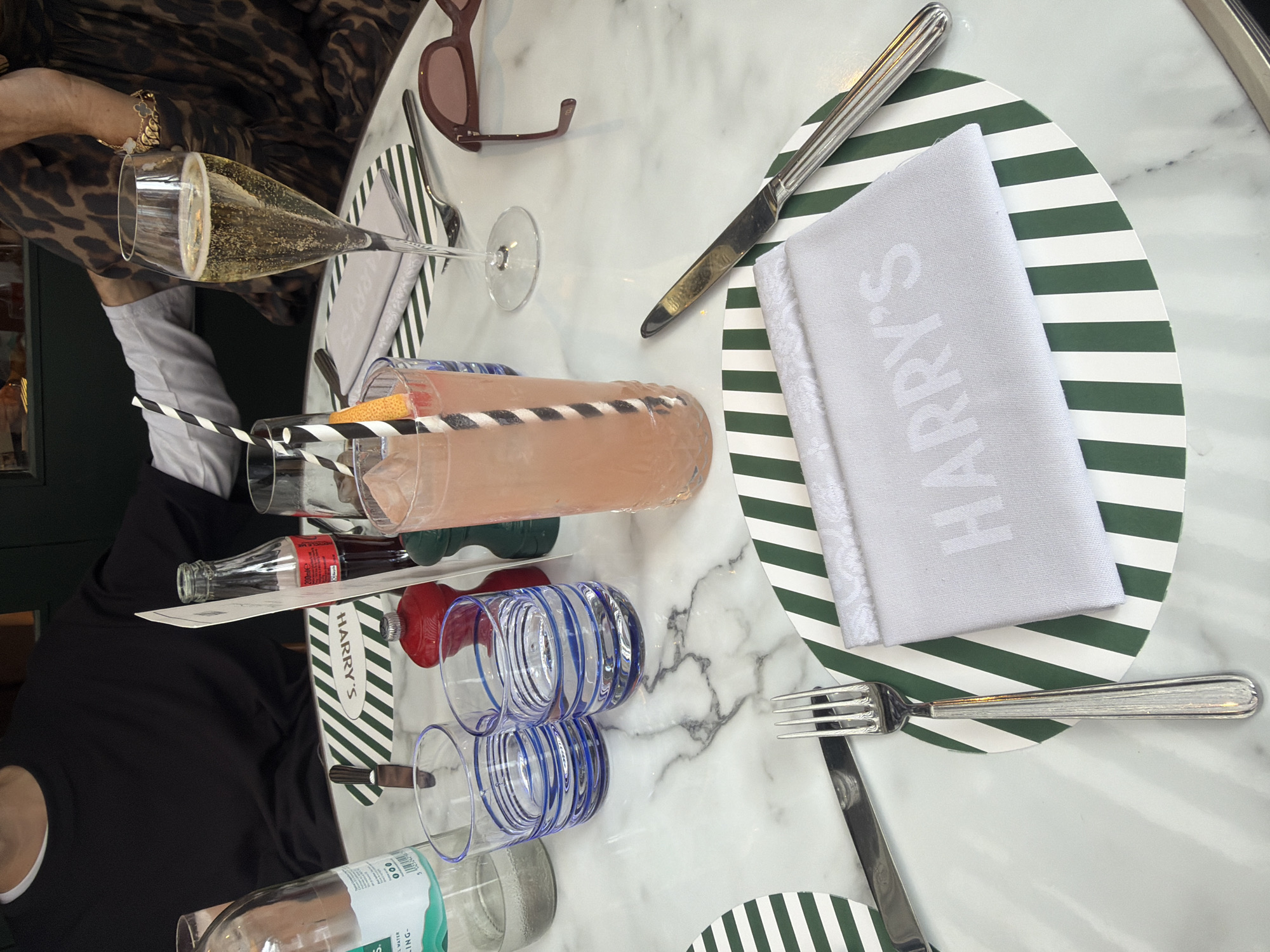 The image shows a table setting at Harry's Bar in London, United Kingdom. The table is covered with a white marble tablecloth. There are two place settings visible, each with a green and white striped placemat.
One place setting includes a white cloth napkin with "HARRY'S" embossed on it, a fork, and a knife. The other place setting, partially visible, also has a green and white striped placemat.
On the table are several drinks. A tall glass contains a pinkish beverage with a striped straw and a piece of citrus garnish. A champagne flute is filled with a sparkling golden beverage. A bottle of Coca-Cola and a bottle of sparkling water are also present.
A pair of sunglasses rests on the table near the champagne flute. Two small salt and pepper shakers are visible between drinks. A person in a black long-sleeved shirt and white undershirt is seated at the table, with their arm resting on the table. Another person, wearing a brown leopard-print dress, is also seated at the table, with their arm adorned with a bracelet resting on the table. The background shows a dark green wall.