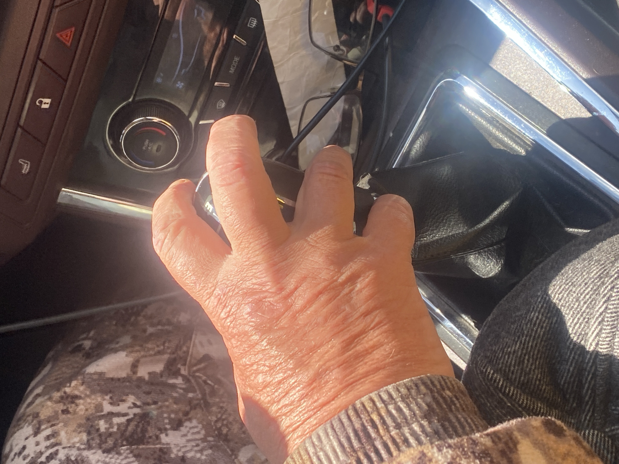 A right hand is gripping a knob on a dark console in a vehicle. The interior of the vehicle is visible, including sections of the dashboard and the passenger seat. The individual is wearing camouflage pants and a sweater with a brown and tan pattern. The image provides no information about the location in Novokuznetsk, Russia.