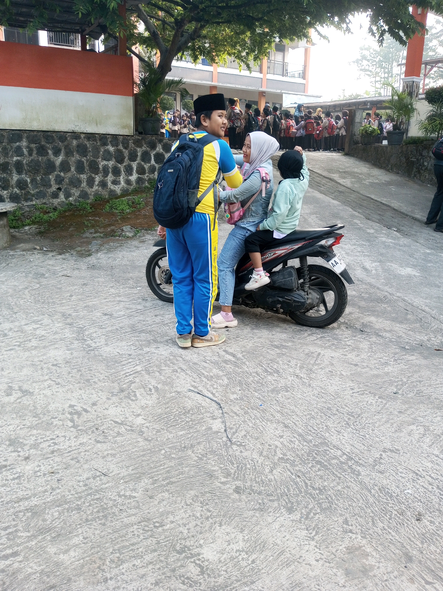 In Krasak, Indonesia, a young boy wearing a blue and yellow uniform and a black peci hat stands next to a motorcycle. He has a black backpack on. A girl in a light blue denim jacket and white hijab is sitting on the motorcycle, with a younger child in a light green top and black hijab sitting behind her. The younger child is reaching out. The motorcycle is black and red. In the background, a line of students in similar uniforms with backpacks are waiting. The scene appears to be at a school. The ground is concrete.