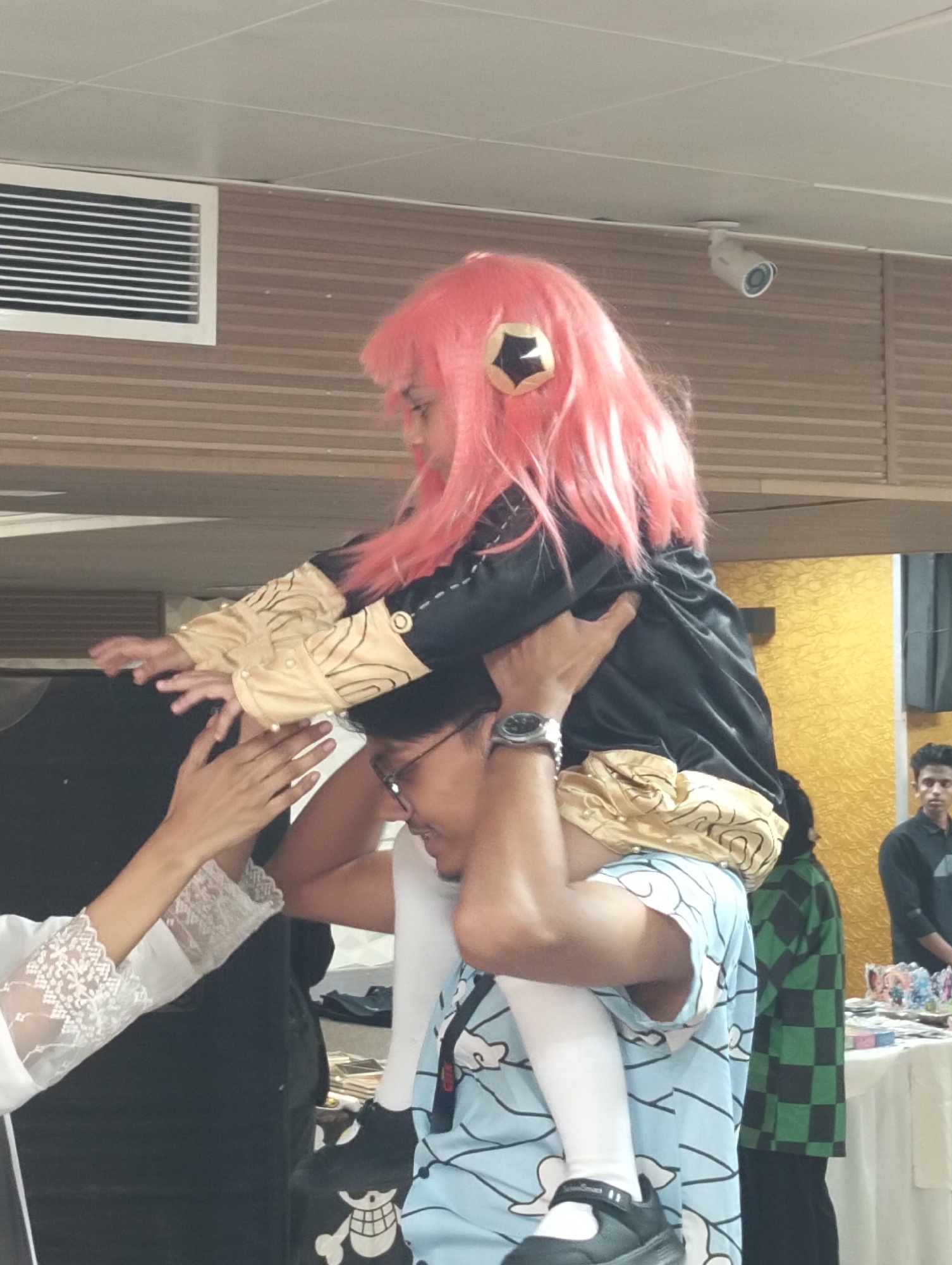 In a room with a drop ceiling and wood-paneled walls, a person is giving a piggyback ride to a child dressed in a costume. The child has bright pink hair and wears a black outfit with gold accents. The person giving the ride is wearing glasses and a blue t-shirt with a cartoon print. To the left, a person in a white lace sleeve is reaching up. In the background, a person in a green checkered outfit stands near a table laden with items. Another person is visible standing further back on the right. A security camera is mounted on the ceiling. This image was taken in Dhaka, Bangladesh.
