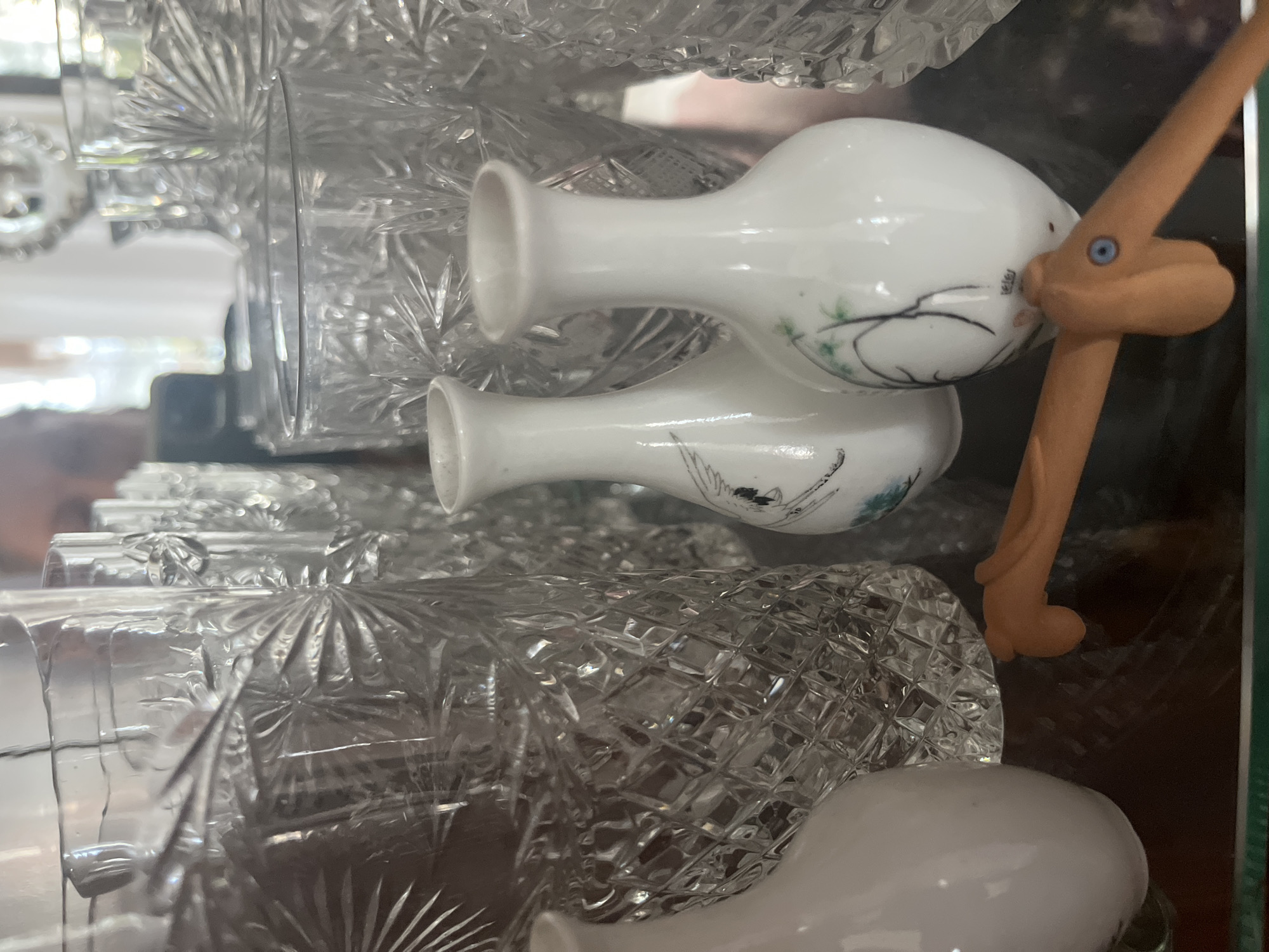 The image shows a close-up, eye-level view of a glass shelf displaying various items. In the foreground, a white, egg-shaped vase with a narrow neck is on the left, and two smaller white vases decorated with bird and branch motifs are positioned towards the center. To the right of these, a portion of a clear, intricately patterned glass vase is visible. Behind these, a row of tall, clear crystal glasses with a starburst pattern on their surface is lined up. In the bottom center, a small, orange, plastic toy figure resembling a bird with blue eyes is placed. The background is out of focus, showing hints of other glassware and a bright, possibly windowed, area. The overall scene is a still life arrangement on a shelf, likely in a domestic setting. The location of Vladimirovka, Russia, is not visually represented in the image.