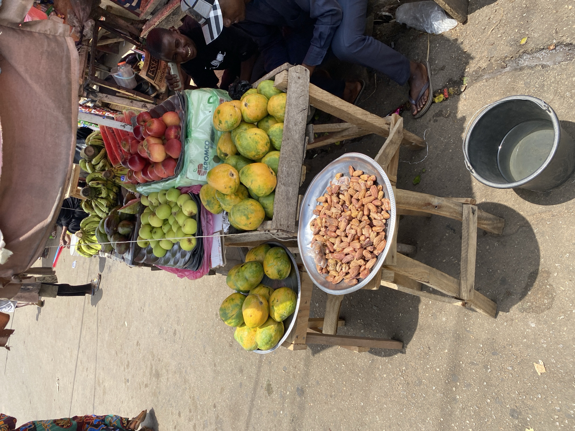 Captured from an elevated angle on a sunlit street in what can be discerned as Jos, Nigeria, the image showcases a bustling fruit stand. A vendor, dressed in black, is visible, interacting with customers around a wooden fruit stand. A person wearing flip-flops is visible near the stand.
The stand is laden with an assortment of fruits: papayas, apples, bananas, and green apples, each neatly arranged for sale. A metal tray sits prominently on the stand, filled with dates. A nearby bucket contains water. The scene is set against a backdrop of street activity.