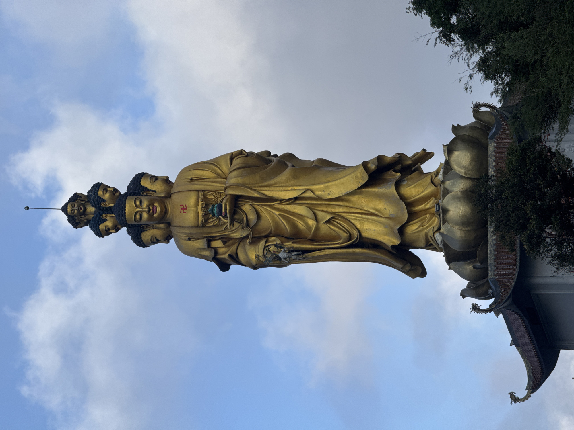 A low-angle, medium shot shows a golden statue with multiple faces, a symbol on its chest, and standing on a lotus flower base, with trees and a traditional building roof to the right and a blue sky with white clouds in the background. The statue is located in Binh Lu, Vietnam.