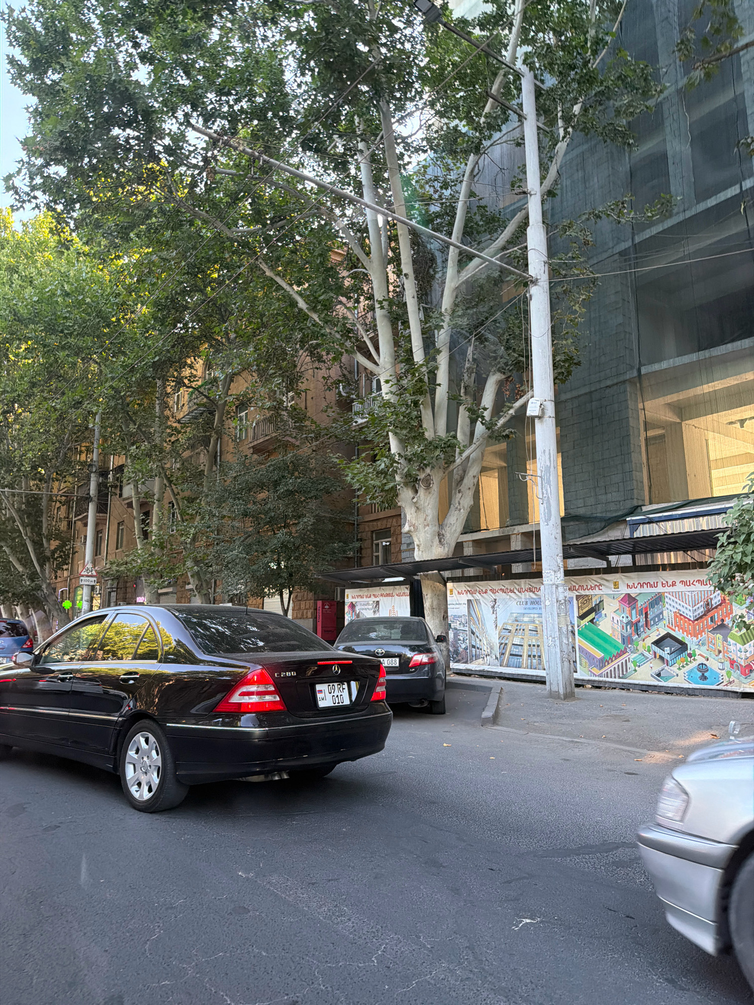 A street scene in Yerevan, Armenia, depicts a black Mercedes-Benz C280 sedan parked on the right side of the road. The car's license plate reads "09 RF 010". Behind it, a silver sedan is partially visible in the foreground. The background shows an older, multi-story building with balconies and windows, partially obscured by large trees with light-colored trunks and green foliage. Further in the background, a building under construction is covered with a dark mesh netting, revealing glimpses of windows with warm lighting. Banners with colorful illustrations and Armenian text are displayed along the sidewalk. The sky is bright, suggesting daylight.