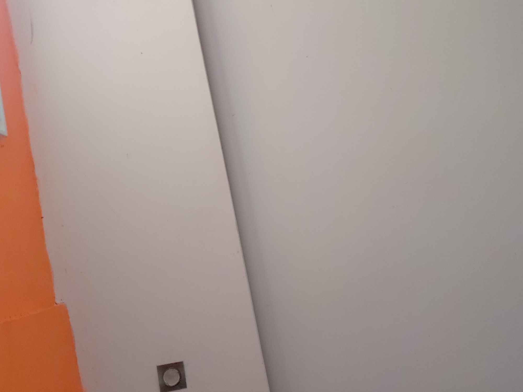The image captures an indoor architectural detail, primarily featuring two intersecting walls or surfaces. The left side of the frame displays a section of a wall painted in a vibrant, uniform orange hue, indicating either an accent wall or an area under renovation. This orange painted surface meets another wall or panel, which is a plain, light gray or off-white color. A sharp, diagonal line forms the boundary between these two dominant surfaces, suggesting a corner or a deliberate architectural joint.
Near the bottom left corner of the frame, on the light gray/off-white surface, there is a small, square metallic object with a circular indentation in its center. This object resembles a flush-mounted electrical outlet, a small light fixture, or a sensor. The overall scene is devoid of people or specific activities, presenting a minimalist and somewhat abstract view of an interior space. The lighting is even, suggesting an indoor environment, possibly with artificial light or diffused natural light. No text is visible. The image conveys a clean, modern aesthetic or a work-in-progress state within a building.