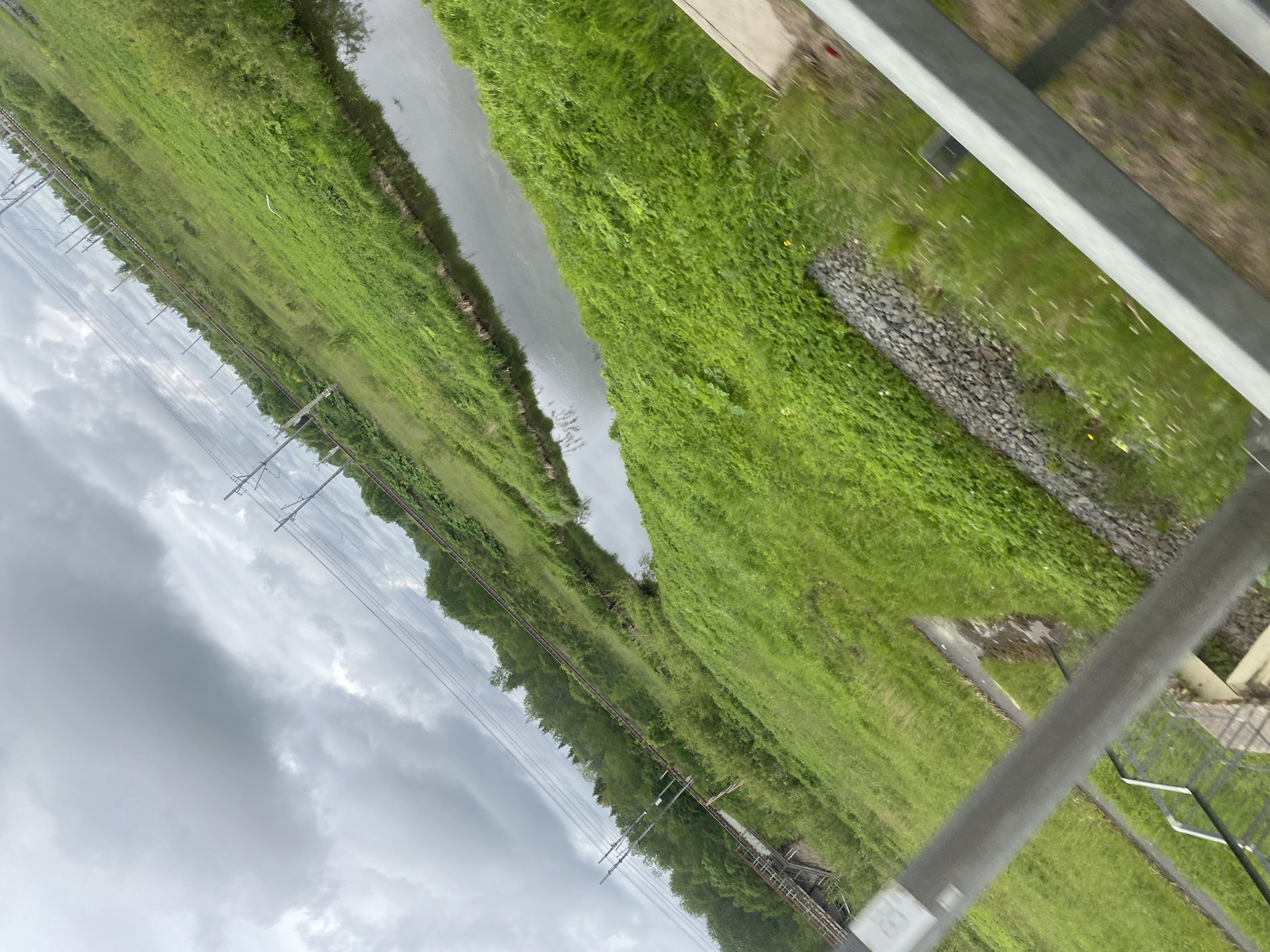 The image presents a view from an elevated position, possibly a train or bridge, overlooking a canal or narrow river. The location is assumed to be near Kommunar, Russia due to the presence of infrastructure typical of that area. The water body is flanked by dense green vegetation, predominantly grass and bushes. Metal railings, likely part of the structure from which the image was taken, are visible in the foreground. The sky is overcast with heavy clouds. Power lines with supporting pylons run along the far side of the water feature. There are no people visible in the image. The overall impression is of a rural or semi-rural scene viewed from a railway or similar infrastructure.