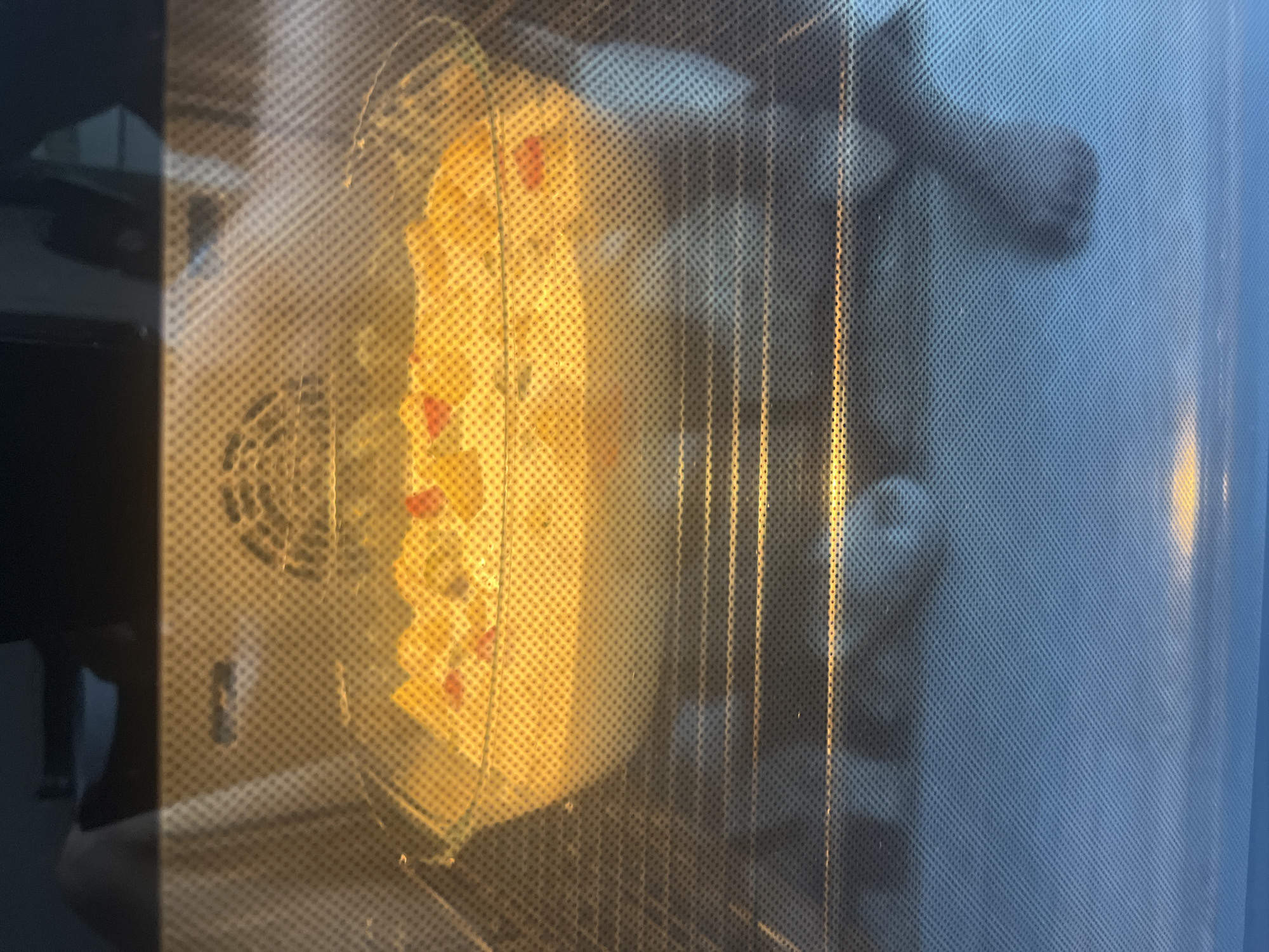 The image depicts a close-up view through the glass door of an oven. Inside, a clear glass baking dish contains food that appears to be a gratin or casserole, with visible pieces of yellow and red ingredients submerged in a creamy sauce. The food is illuminated by the oven's interior light, casting a warm glow. The oven racks are visible below the dish. Reflections on the oven door suggest the presence of ambient light and potentially furniture in the surrounding environment. The image does not contain any specific visual cues that would allow for the determination of its location in Burgas, Bulgaria.
