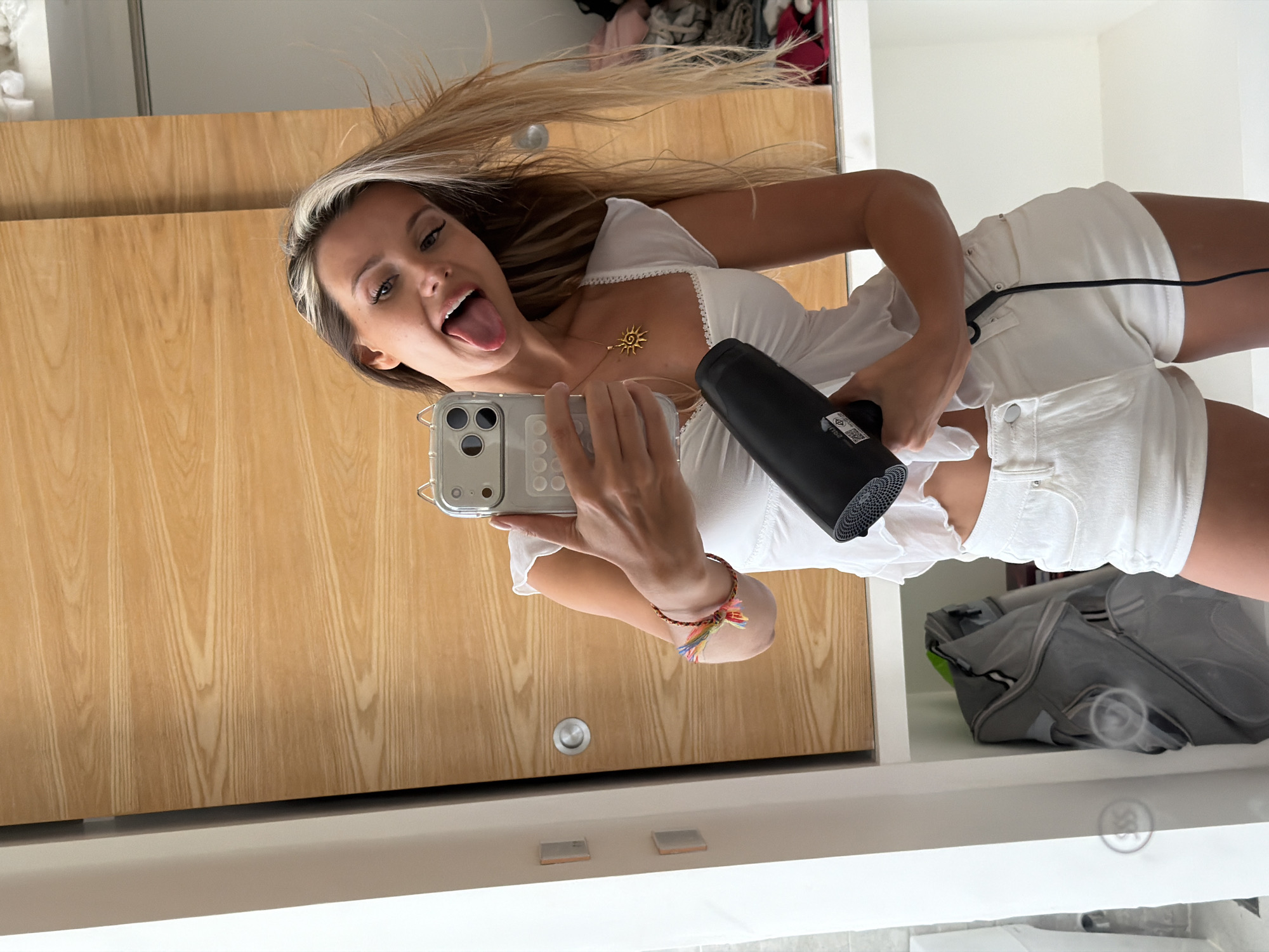 The image shows a young woman with blonde hair, wearing a white top and white shorts, holding a black hairdryer and a smartphone. She is sticking her tongue out and looking at the phone's camera, which is positioned as if to take a selfie. The background features a wooden wardrobe with sliding doors and a white shelf. A grey backpack is visible on the shelf below the woman. The overall setting appears to be an indoor space, possibly a room or a closet, and there is no indication of it being located in Thep Krasatti, Thailand.