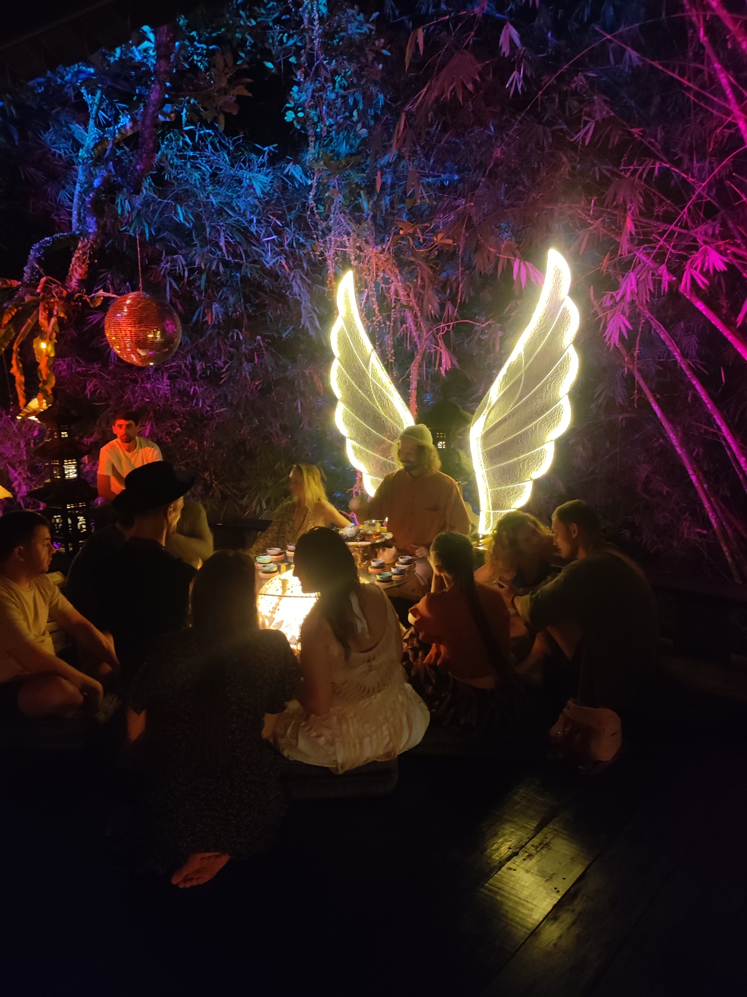 The image depicts a nighttime gathering of approximately ten people seated in a circle on a wooden floor in an outdoor setting in Lokaserena, Indonesia. A man with long hair and a light-colored shirt appears to be serving or presiding over a tea ceremony, with small cups arranged on a table. Behind him are illuminated angel wings, acting as a focal point. The environment is decorated with bamboo and other foliage, lit with blue and purple lighting. A disco ball hangs from a tree above on the left, and a man sits in the background on the left.