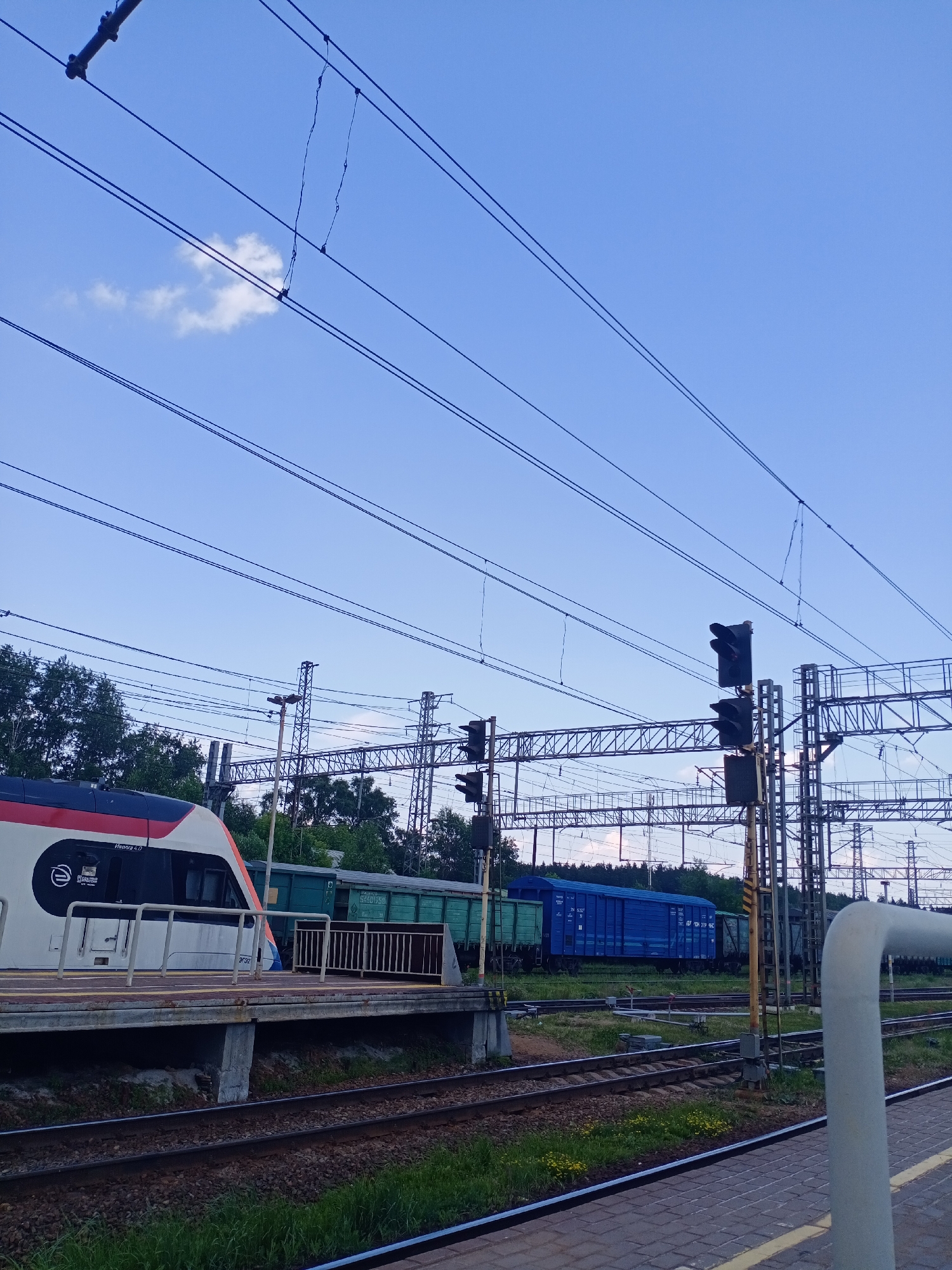 A train is at a station in Fryazevo, Russia. The train is white with red and blue accents. It is on tracks at a platform. In the background, there are other trains, some green and some blue. There are also trees, power lines, and a traffic signal. The sky is blue with a few white clouds.