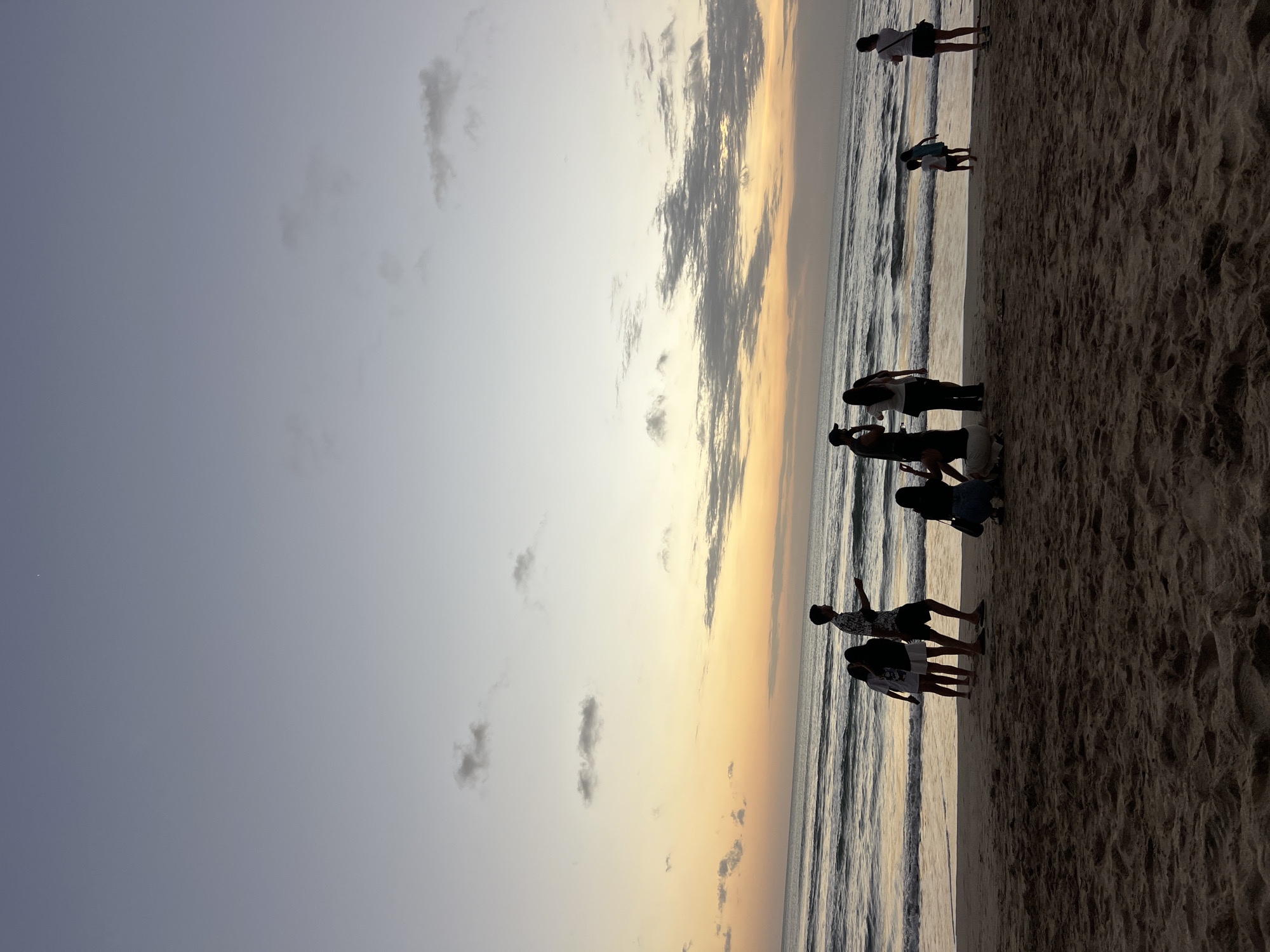 A group of people are gathered on the sandy beach of Kuta, Indonesia, near the water's edge. The sky above is a gradient of pale blue and soft orange, indicating either sunrise or sunset. The ocean waves are gently rolling onto the shore. Silhouettes of individuals and small groups are visible, some standing and others appearing to be sitting or crouching. The scene appears to be a casual gathering, possibly a family or friends enjoying the coastal environment. The sand in the foreground is textured and appears dry.