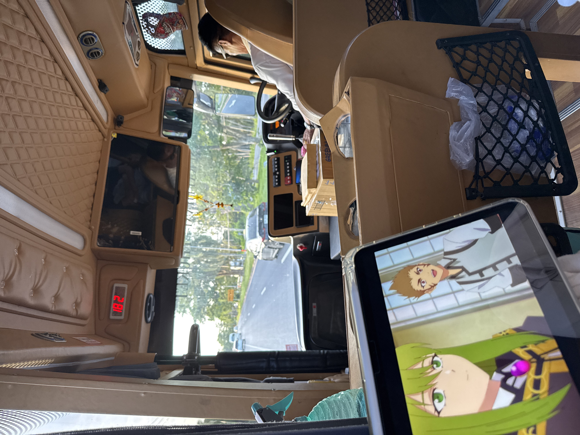 The image shows the interior of a bus, with a driver in the foreground. The bus is traveling on a road in Ban Suanma Prao, Thailand. The driver is a man with glasses and short hair, wearing a white shirt. He is holding the steering wheel and looking at the road. The bus has a tablet in the foreground, displaying an anime. The tablet shows two characters, one with green hair and the other with brown hair. The bus has comfortable seating and interior lighting. There are also storage compartments and a spray bottle visible. The road outside is lined with trees and other vehicles.