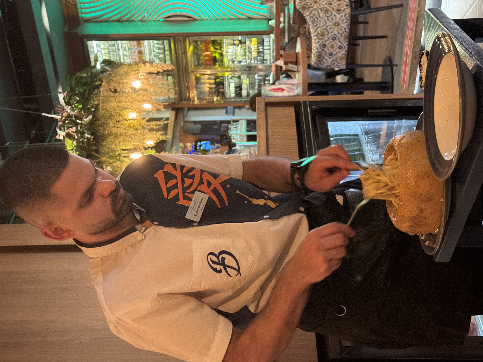 A male server, identified by a name tag as "Валентин Митов" from "Burrata" and "BAMBOO," is meticulously serving pasta in a restaurant setting in Sofia, Bulgaria. He has short dark hair, a beard, and wears a distinctive short-sleeved shirt that is half white (left side) with a dark embroidered "B" on the pocket, and half dark blue (right side) featuring orange characters and white/yellow paint splatters. He also wears a black apron and a dark wristwatch on his left wrist. Using a fork and spoon, he is actively transferring spaghetti-like pasta from a large, hollowed-out bread bowl, resting on a silver tray, into a dark blue-grey ceramic bowl containing a creamy, light-colored sauce. A small dark bowl holding a white condiment is positioned behind the serving bowl. The environment is a dimly lit restaurant interior, featuring a hanging plant decoration with small string lights. In the background, refrigerated display cases are visible, stocked with numerous bottles. To the right, a prominent decorative wall panel with vertical wavy lines emits a vibrant green light, and patterned restaurant chairs are visible further back.