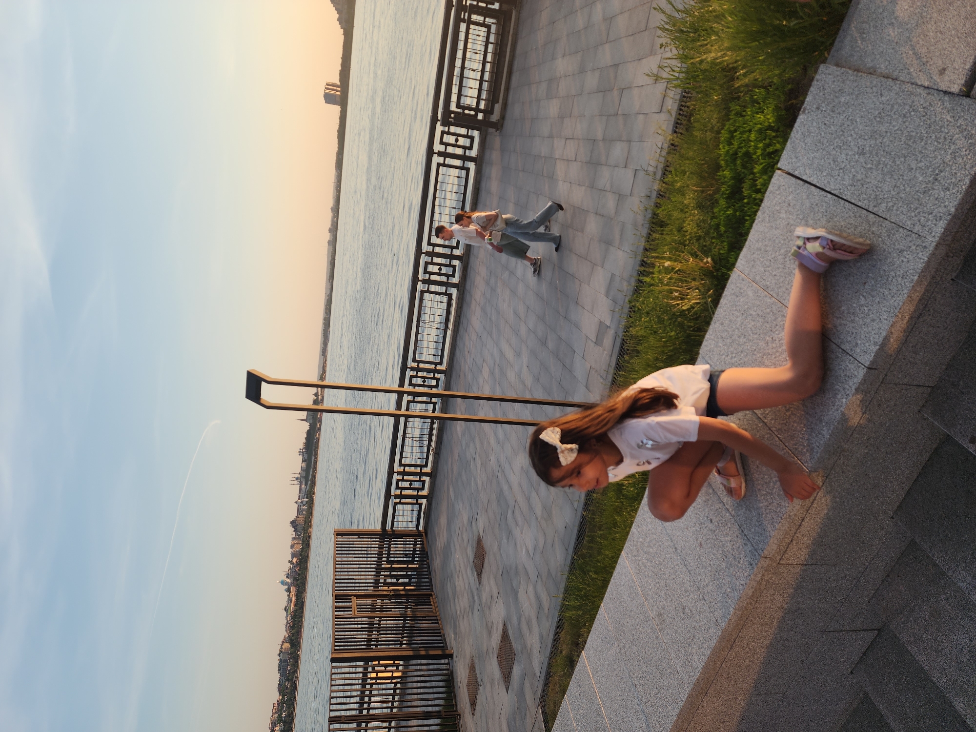 The image depicts a young girl at an outdoor location in Unknown, Unknown. She is positioned on a stone structure, possibly a retaining wall or tiered ledge. The girl is fair-skinned with brown hair and a white bow. She is wearing a white t-shirt and dark shorts and pink sandals. She is positioned in a crouched position.
Behind her, there is a waterfront walkway featuring a metal railing, and a couple is seen walking in the background. The landscape includes a body of water, and the sky appears to be at sunset. The buildings are across the water. There is some greenery on the walkway.