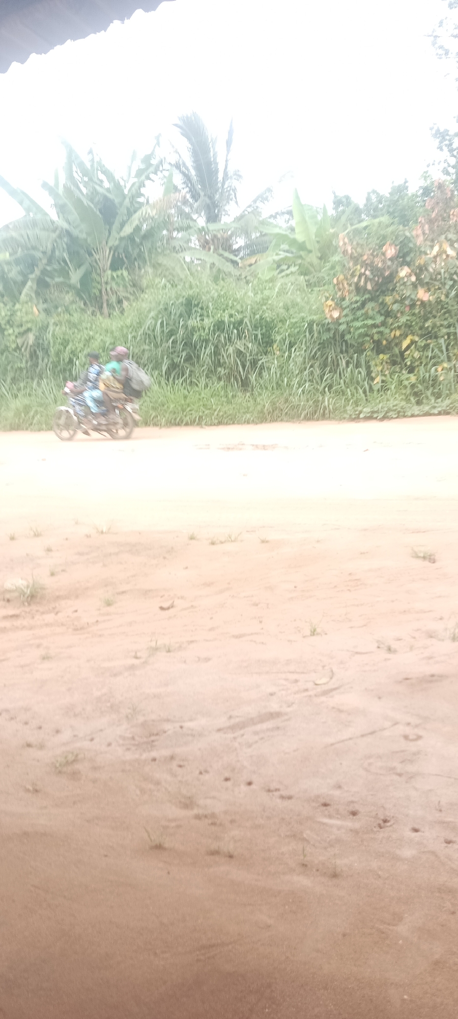 The image shows a dirt road in Amauzu, Nigeria. A motorcycle with two people on it is riding along the road. The road is surrounded by tall grass and trees.