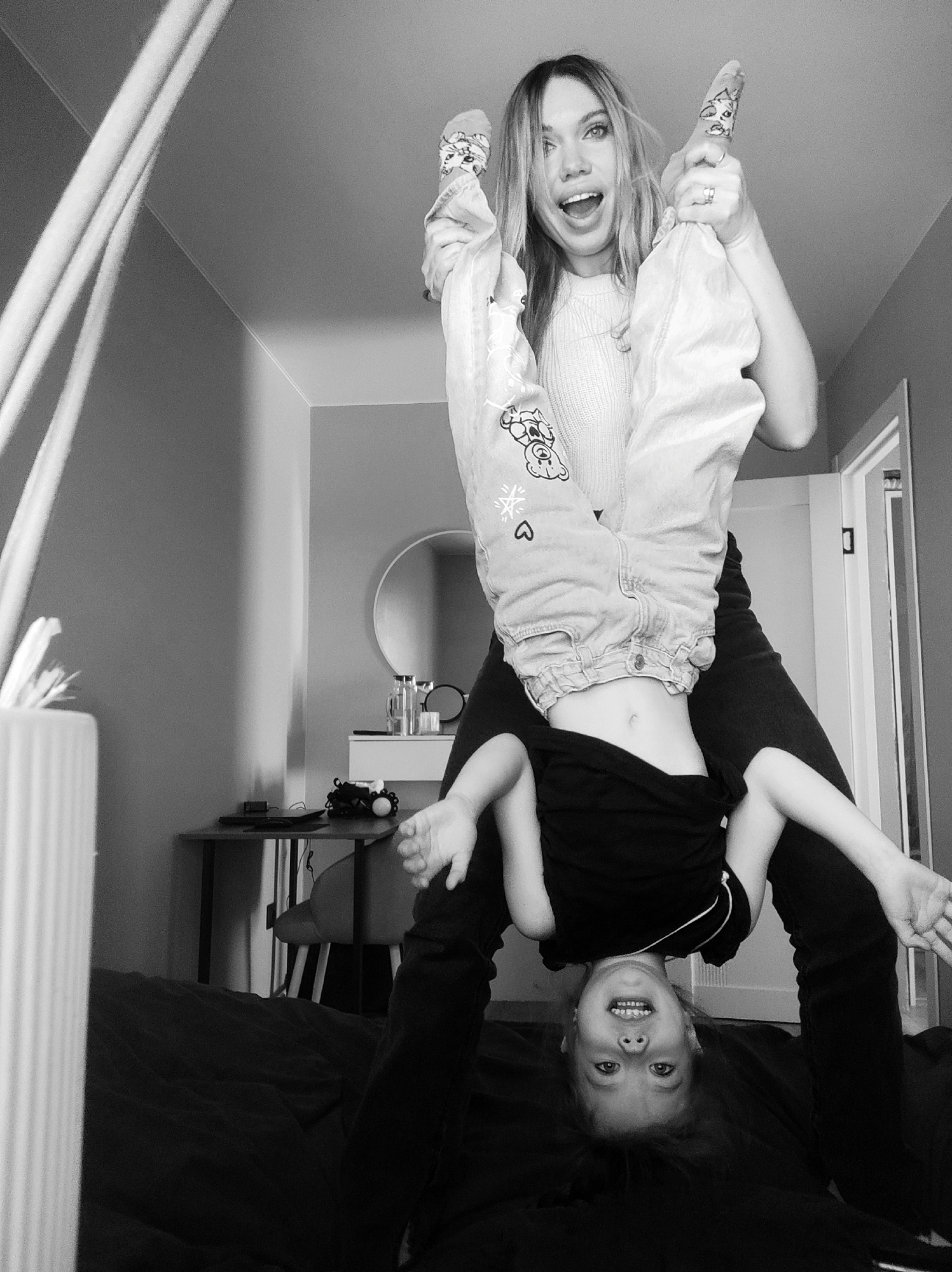 A black and white medium shot depicts a woman holding a child upside down indoors. The woman has long hair and is holding the child's feet, grinning. The child, dressed in a dark shirt, is also smiling with arms outstretched, upside down with the head close to the floor. Behind the woman, a desk is visible with objects including a mirror and table. The photo is taken indoors, likely a bedroom. There is no indication this image was taken in Votkinsk, Russia.