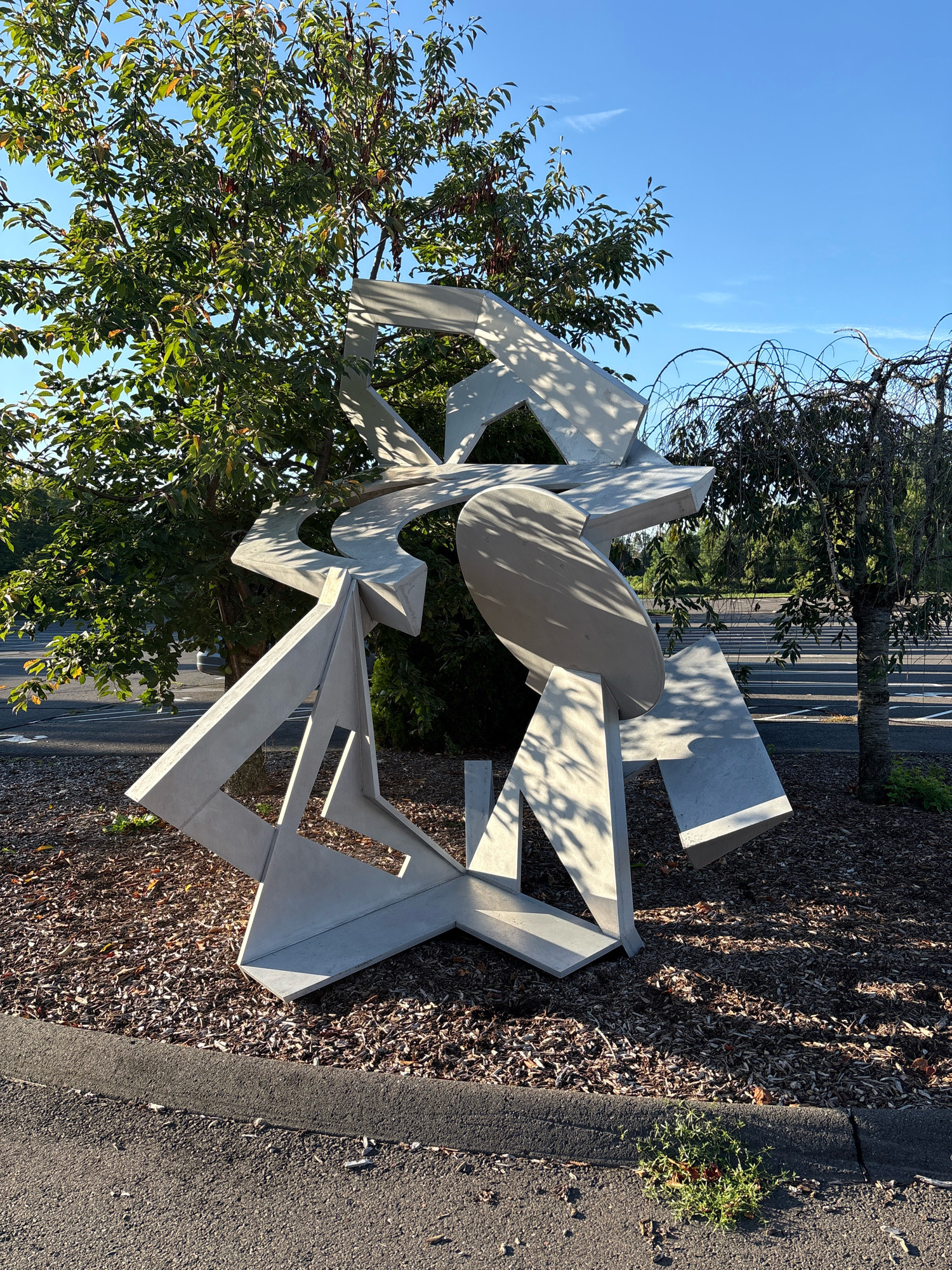 A contemporary abstract sculpture made of concrete sits outdoors on a bed of wood chips. The sculpture is composed of interlocking geometric shapes, primarily sharp angles and planes, with a large elliptical element in the center. The piece casts distinct shadows on itself and the surrounding ground, indicating direct sunlight. Behind the sculpture, a leafy green tree with dappled sunlight filtering through its branches dominates the left side of the frame. To the right, a sparser, weeping tree with dark branches is visible, extending towards the right edge. In the background, beyond the trees, is a paved area, likely a parking lot, with faint white lines visible on the pavement. Further in the distance, a line of trees forms a dense green backdrop under a clear blue sky with a few wispy clouds. The foreground shows a paved asphalt surface with a darker gray curbing, and a small patch of green vegetation growing between the curb and the asphalt. The overall setting appears to be a public outdoor space.