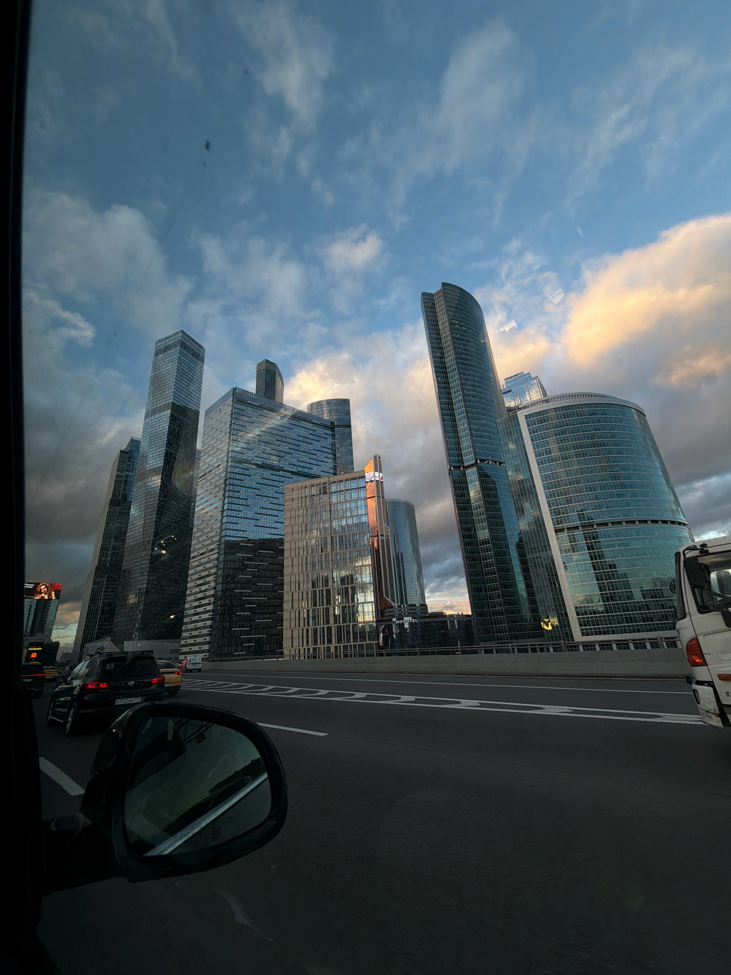 A low-angle, wide shot captures the Moscow International Business Center, also known as Moscow City, on a cloudy day. The skyscrapers, characterized by their modern glass facades, dominate the skyline. In the foreground, a dark-colored car is seen from the driver's perspective, with a side-view mirror reflecting the sky and buildings. A highway runs in front of the skyscrapers, with other vehicles, including a black SUV with a license plate that appears to read "0728GO77", and a white truck, traveling on it. The sky is filled with dramatic clouds, with patches of sunlight breaking through, illuminating some of the buildings and the clouds. The overall impression is one of urban grandeur and movement.