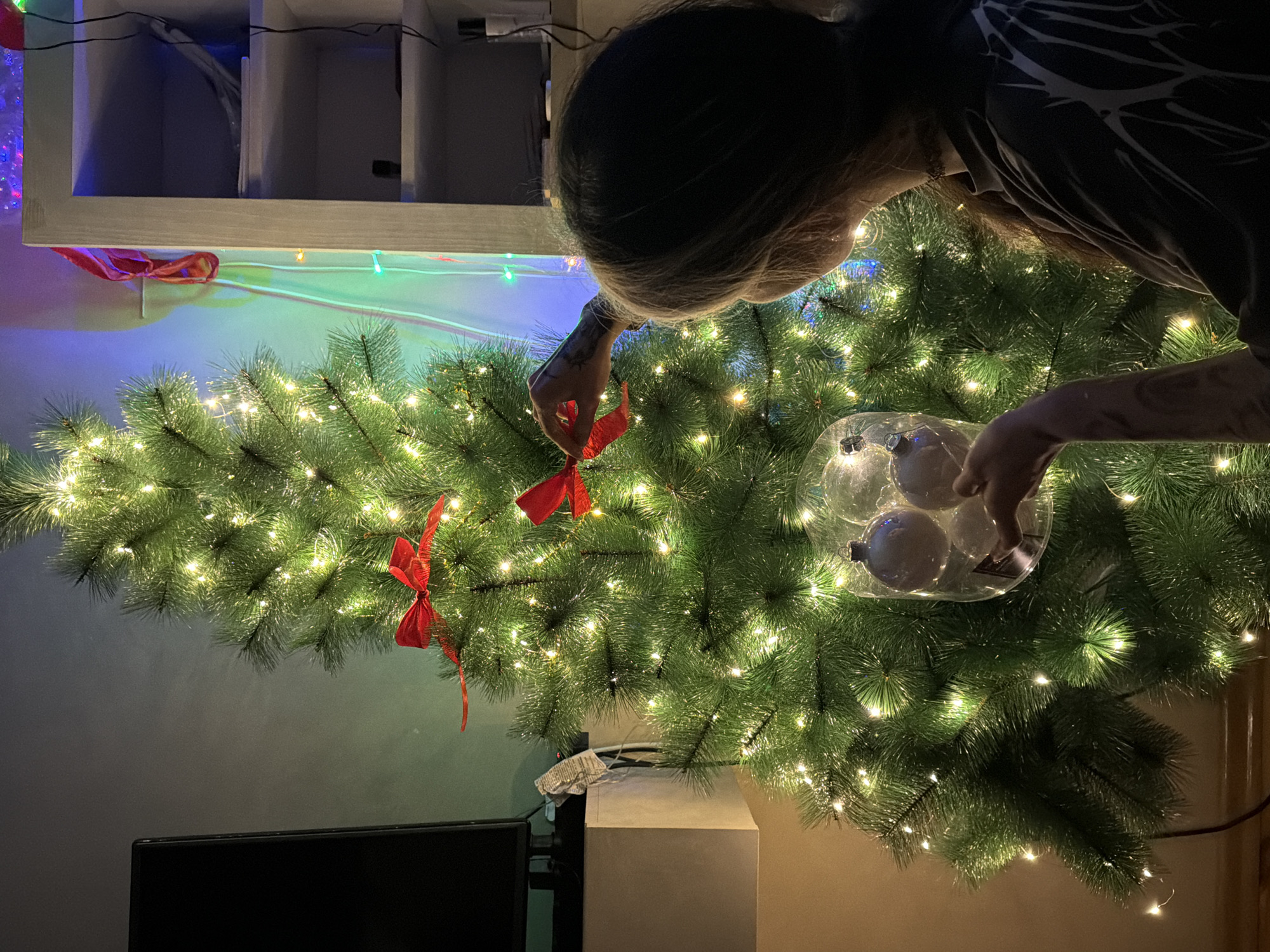 The scene depicts an interior room located in Budva, Montenegro. One individual, viewed from behind and to the right, is engaged in decorating a Christmas tree. The person has long, light-brown hair and is wearing a dark, short-sleeved top with a white, branching pattern on the back. A choker necklace is visible. The individual's right forearm displays a dark tattoo.
The primary subject is a green artificial Christmas tree, brightly illuminated with numerous small, warm white LED lights. Several red satin bows are visible among the branches, with the person's right hand adjusting one near the upper-middle section of the tree. The person's left hand is holding a transparent plastic container that holds three spherical ornaments: two are clear or frosted white, and one is pale blue or silver-grey.
In the background to the left, a dark flat-screen television is visible, resting on a light-colored stand or cabinet. Wires are present behind the television. To the right, a white open shelving unit with multiple shelves is situated. Indistinct items occupy these shelves, including a dark cylindrical object on one shelf. The wall behind the tree and shelving unit displays ambient colorful lighting, suggesting additional decorative light sources. The room is dimly lit, emphasizing the glowing lights of the Christmas tree and other decorations.