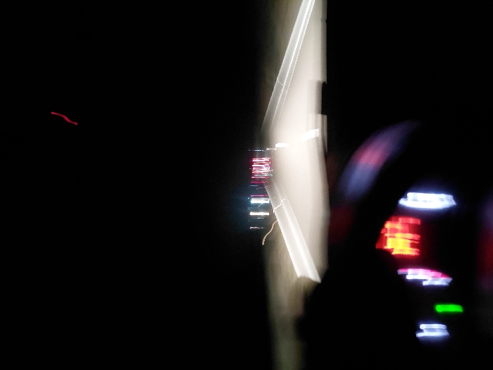 A blurry, long-exposure shot captures a nighttime scene from inside a moving vehicle. A dashboard with illuminated gauges glows on the right. The road ahead is visible as a bright streak of light with faint hints of oncoming headlights and taillights. The left side of the image is predominantly dark, showing a faint red streak. The motion-blurred lights suggest the vehicle is traveling at a significant speed.