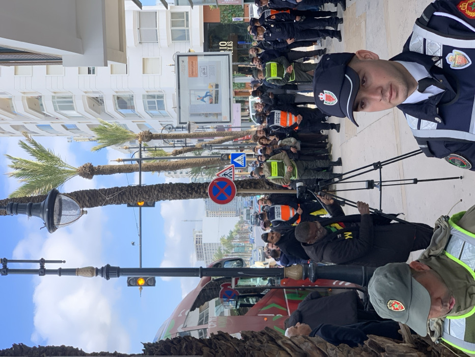 The image displays an urban street scene in Tangier, Morocco, characterized by a significant presence of law enforcement and a public gathering.
In the immediate foreground, a male individual, identifiable as a police officer by his dark blue uniform, reflective stripes, and peaked cap bearing a Moroccan royal crest, is positioned on the right, looking directly forward. To his left, partially visible, is another individual in a green reflective vest and matching cap, also associated with law enforcement.
In the midground, a dark-skinned man with glasses and a beard is actively adjusting a camera mounted on a black tripod on the sidewalk. He wears a dark jacket over a multi-colored shirt featuring green, yellow, red, and the letter 'W'. Behind him and extending to the right, a crowd of diverse individuals, predominantly male, is gathered on the wide paved sidewalk. Several uniformed police officers, wearing dark blue uniforms with orange or green reflective vests marked "POLICE" and "الشرطة" (Arabic for Police), are interspersed within and at the periphery of the crowd, indicating a security or crowd management function. A large red and green bus is partially visible on the far left, stopped or parked on the street.
The environment includes a multi-lane road extending into the distance with various vehicles. Tall palm trees line the street and central median. Black ornate street lamps and traffic lights displaying yellow signals are prominent. Modern, multi-story white buildings with balconies and commercial ground floors, one displaying "RIO TEXAS," line the street. Street signage includes a "No Stopping" sign and a pedestrian crossing sign. A large, illuminated outdoor advertisement is visible on the right side of the street. The sky above is partly cloudy.
