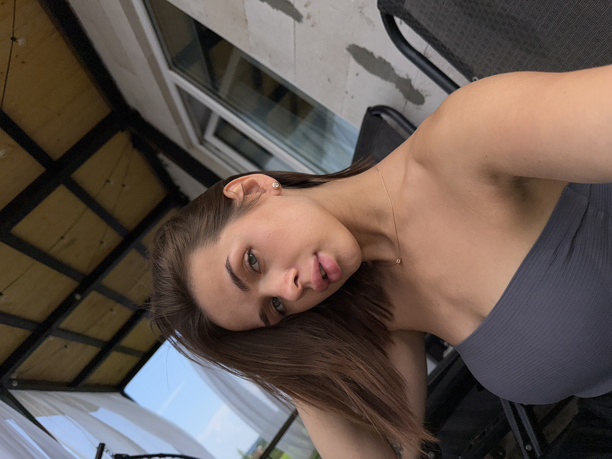 A young woman is posing for a selfie outdoors, with what appears to be a patio or balcony area. She has brown hair, fair skin, and blue eyes. She is wearing a grey strapless top and a delicate gold necklace. Her left hand is resting on her head, and her right arm is extended to take the photo. In the background, a covered structure with wooden beams and translucent white fabric can be seen, along with a window and the side of a building. Dark chairs are visible in the foreground and background. The environment suggests a residential or recreational outdoor space. There is no indication of the specific location within Lobanovo, Russia.