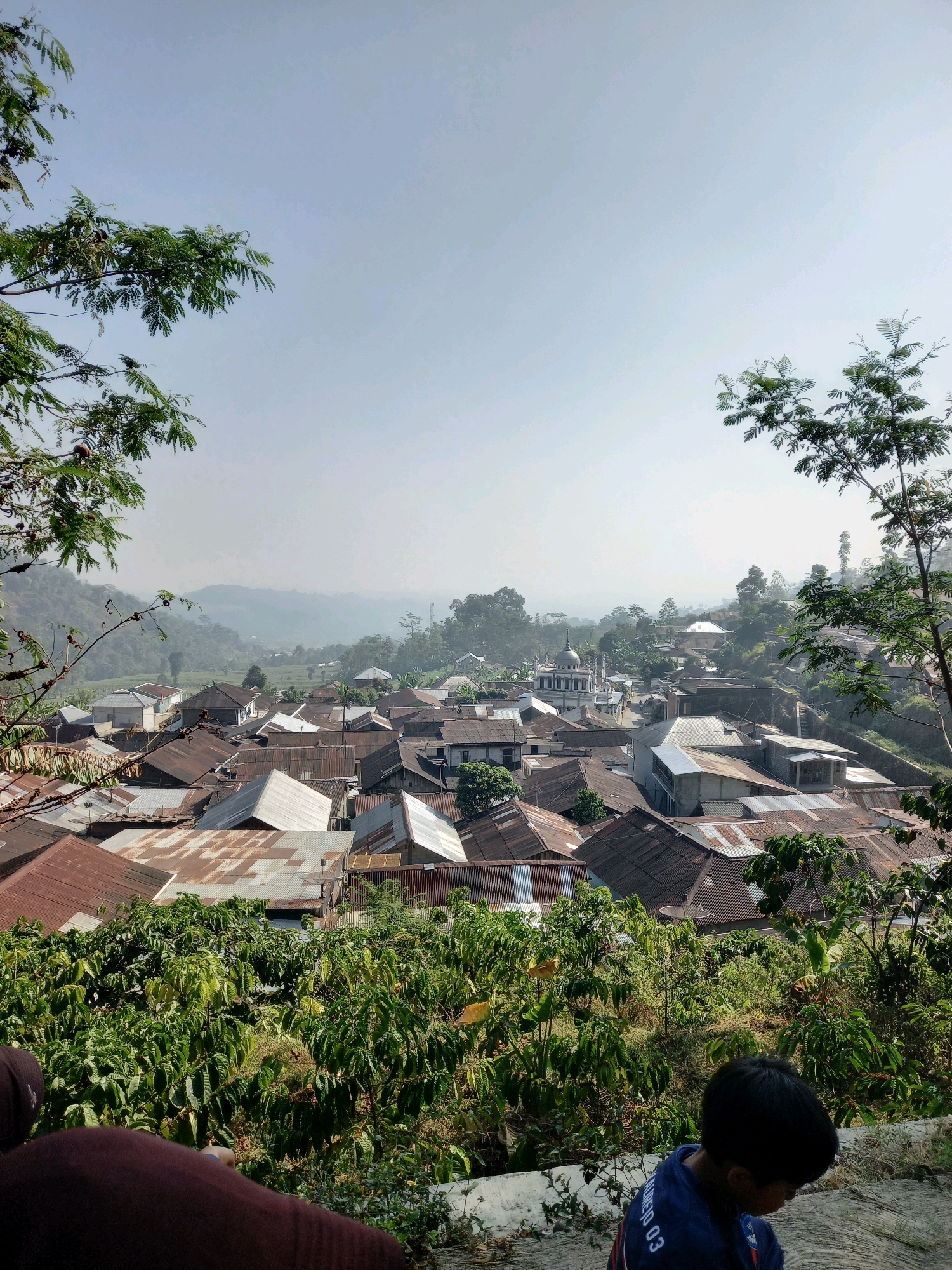 An aerial, high-angle, vertical shot captures a dense village nestled amidst rolling hills under a hazy, pale blue sky. The village is characterized by numerous houses with corrugated metal roofs, many of which appear weathered and rusted, creating a mosaic of earth tones. In the center of the village, a white building with a dome and minaret, possibly a mosque, stands out. Lush green vegetation, including trees and bushes, surrounds and punctuates the settlement, with larger trees framing the scene from the foreground and sides. In the lower right corner, a young Black boy with short hair, wearing a dark blue shirt with white lettering, is partially visible, looking downwards. In the lower left, the top of another person's head and shoulder, clad in dark clothing, are also present, facing away from the camera. The overall impression is of a rural Indonesian community, possibly located in the area of Blumah, Indonesia, seen from an elevated viewpoint.